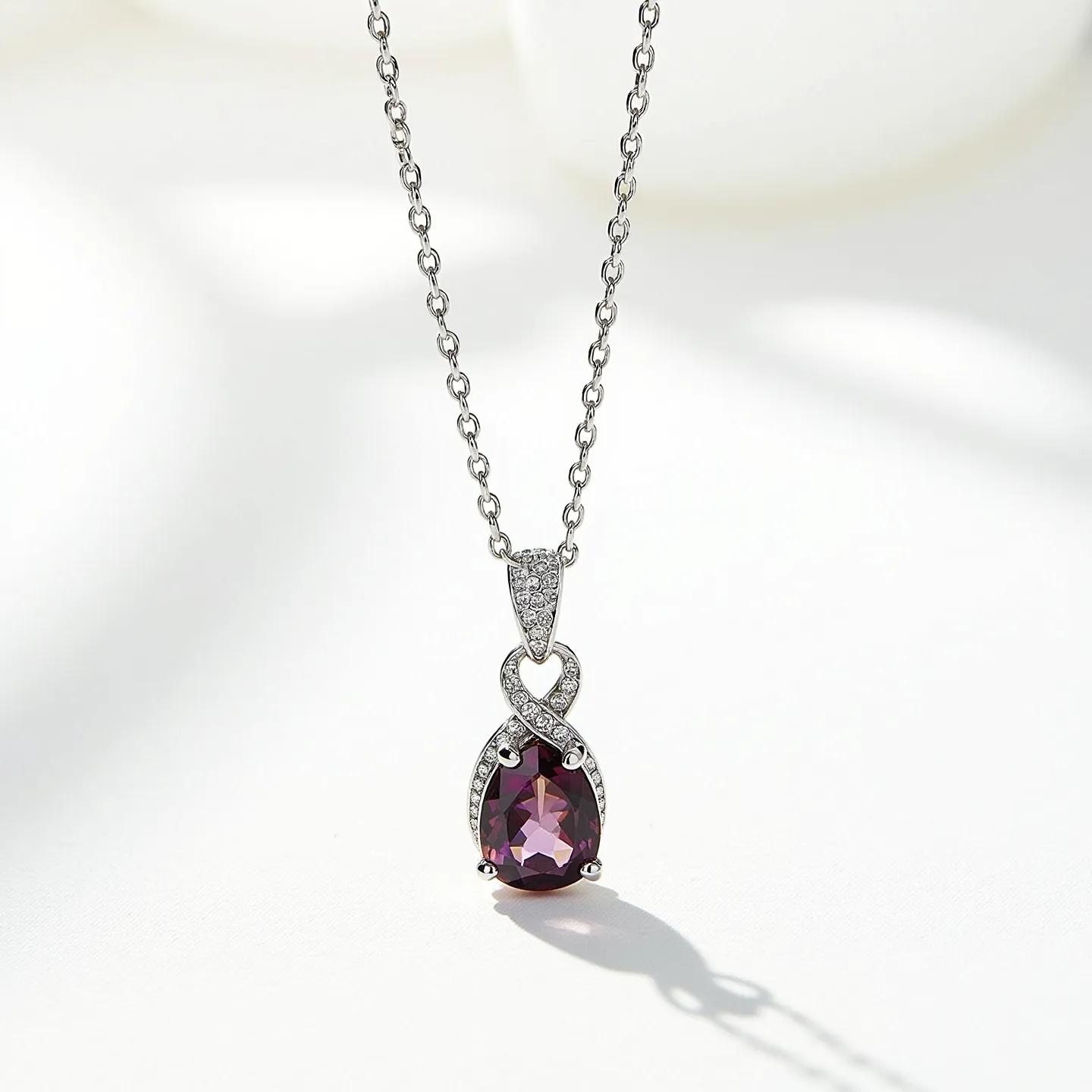 This stone necklace features a striking pendant with a deep purple, teardrop-shaped gemstone, likely amethyst or a similarly colored stone, set in a classic prong setting. The gem is elegantly encased in swirling silver metal, adorned with small, clear stones that resemble diamonds, enhancing its sparkling appeal. The pendant is suspended from a delicate silver or white gold chain with a simple lobster clasp, ensuring a secure fit. The intricate setting adds a touch of sophistication, making it a versatile piece suitable for both casual and formal occasions.