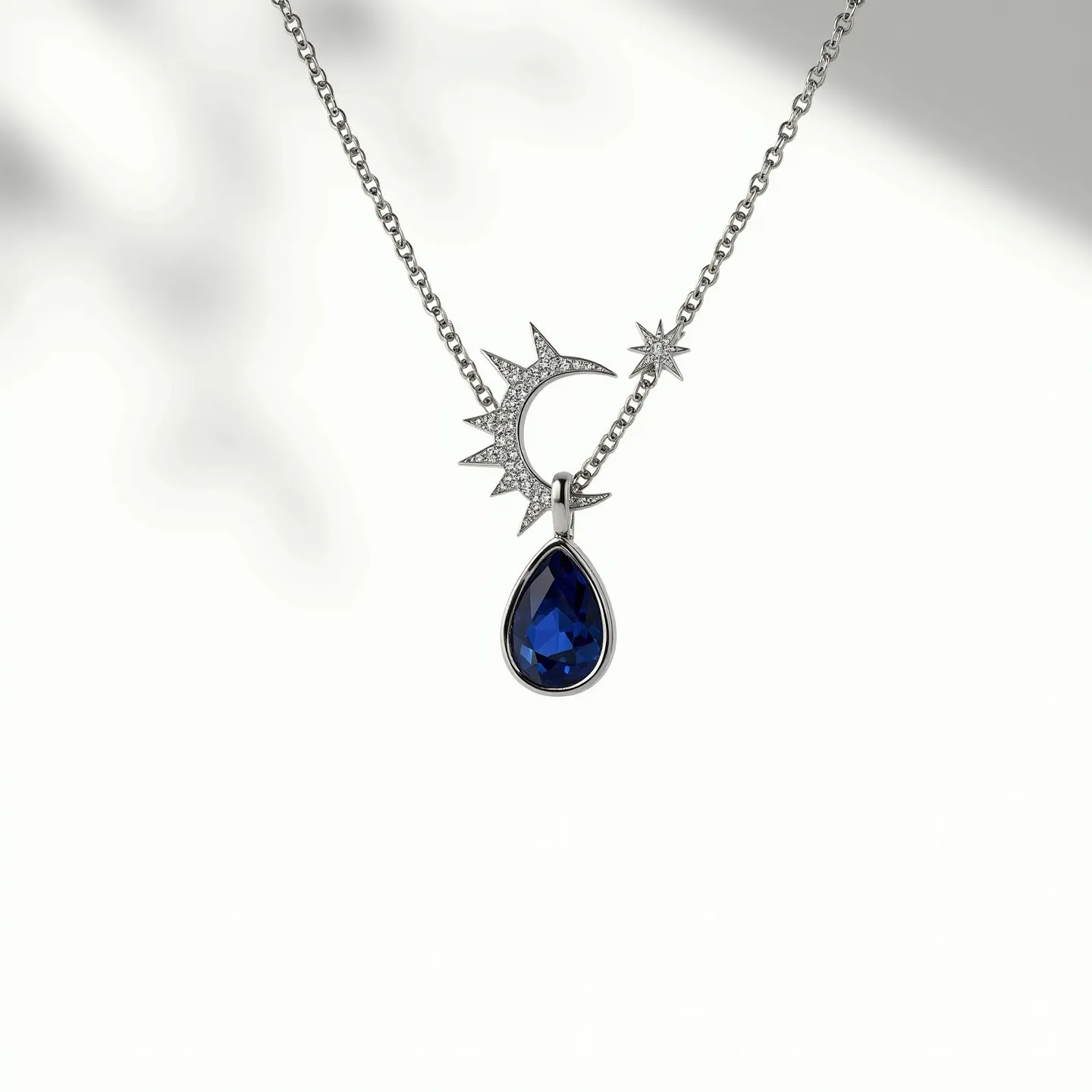 This stone necklace features a delicate silver chain that elegantly supports celestial-themed pendants. Central to the design is a striking teardrop-shaped blue gemstone, possibly a sapphire or blue topaz, set in a simple bezel setting that enhances its deep hue. Adjacent to the gemstone are two decorative elements: a crescent moon adorned with small, sparkling stones that may be diamonds, adding a touch of shimmer, and a small star accentuating the celestial motif. The necklace is completed with a secure clasp ensuring both style and function.