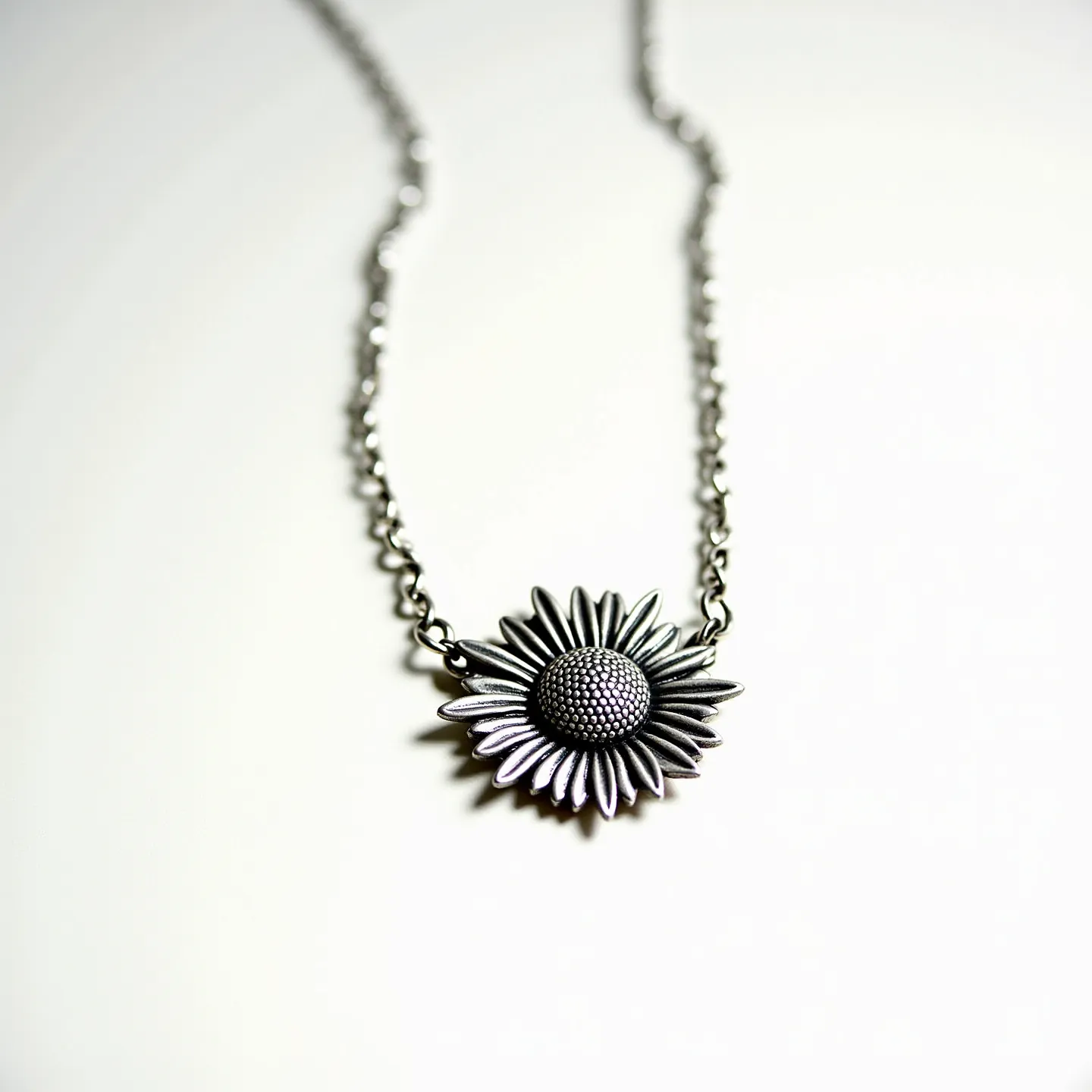 This sunflower necklace features a delicately crafted sunflower pendant made of a metallic material, likely silver or a similar alloy, which is polished to enhance its intricate details. The sunflower's petals are finely textured, adding depth and realism to the design. The central disc of the flower has a textured surface to mimic the appearance of sunflower seeds, further capturing the flower’s natural appearance. The pendant is suspended on a simple chain made from the same metallic material, which complements the floral centerpiece. The necklace is equipped with a standard clasp, ensuring secure wearability while allowing for easy attachment and removal.