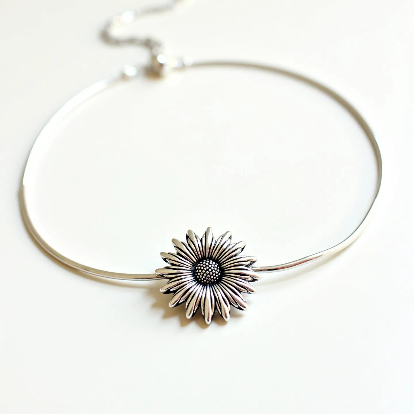 This sunflower necklace features a beautifully crafted sunflower pendant, likely made of silver or a similar metal, with intricately detailed petals surrounding a textured center. The design captures the natural beauty of a sunflower with a modern touch. It appears that there are no additional gemstones embedded in the piece. The necklace is suspended on a sleek choker-style band, possibly made of the same metal as the pendant, providing a seamless and elegant look. The attachment includes a delicate clasp mechanism, likely a lobster clasp, which allows for easy fastening and adjustment to ensure a comfortable fit around the neck.