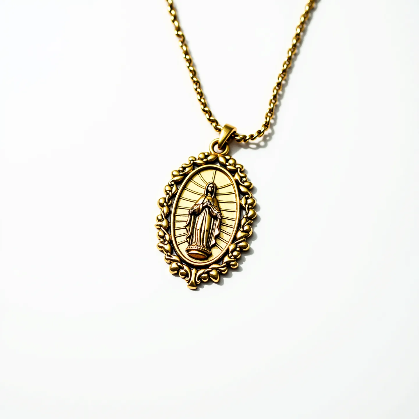 This Virgin Mary necklace features a gold-toned oval pendant with an intricately detailed depiction of the Virgin Mary at its center. The pendant is adorned with a radiant design surrounding the figure, adding depth and dimension to the piece. It is bordered by an ornate frame resembling small floral motifs. The pendant is suspended from a simple yet elegant gold-toned chain, which is designed to complement the pendant’s aesthetic. The necklace appears to have a straightforward link chain design, with a standard clasp that secures the necklace when worn.