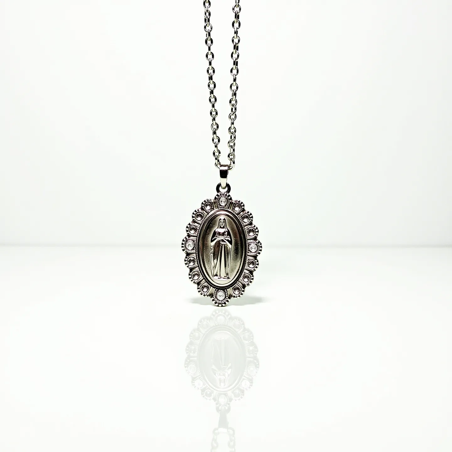 This virgin Mary necklace features an intricately designed pendant, likely made of silver or a silver-toned metal, with an oval shape that frames the depiction of the Virgin Mary at its center. Surrounding the central figure is a detailed border with a series of small, evenly spaced circular embellishments that resemble either beads or metalwork designed to mimic the appearance of gemstones. The pendant is suspended from a simple chain composed of interconnected links, and it appears to have a standard jump ring or bail attachment, allowing for smooth movement along the chain. The overall design combines religious symbolism with decorative artistry, making it both a spiritual and aesthetic piece.