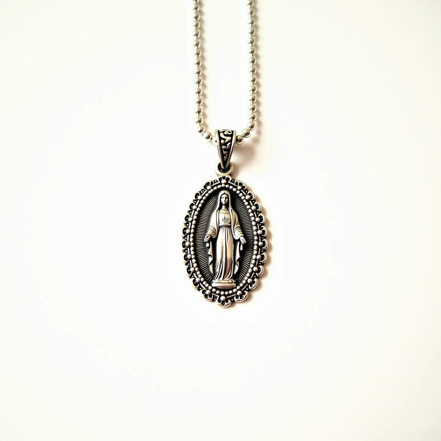 This Virgin Mary necklace features an intricately designed oval pendant made of silver-tone metal, showcasing the figure of the Virgin Mary in a detailed raised relief. The pendant is surrounded by a decorative filigree border that enhances its ornate appearance. It hangs from a ball chain, also silver-toned, which complements the pendant's aesthetic. The necklace is equipped with a standard connector clasp for secure attachment, providing both functionality and style.