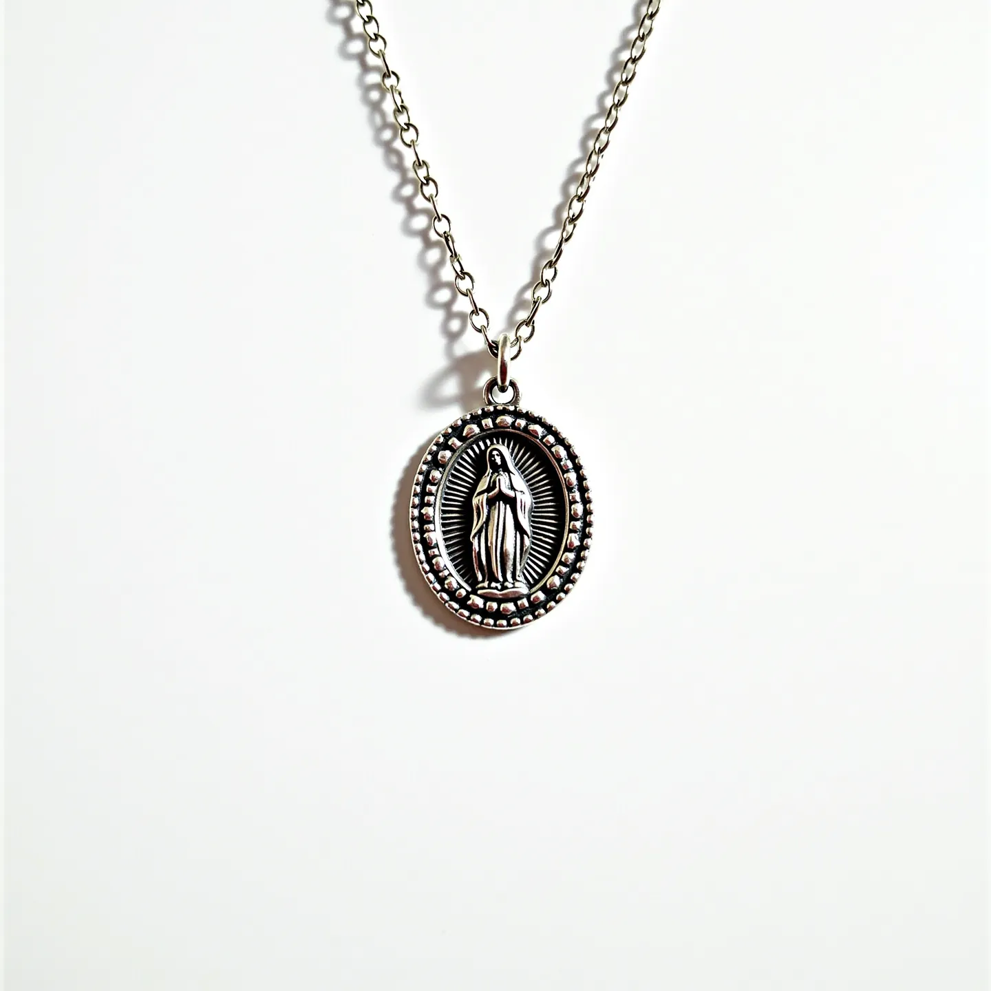 This Virgin Mary necklace features an oval pendant, likely made of silver or a silver-toned metal, depicting the Virgin Mary in a classic pose with engraved details. The pendant is surrounded by a beaded border, adding a texture that emphasizes the central figure. Attached to a simple chain, the necklace appears to have a standard lobster clasp, providing a secure fastening. The overall design is understated yet elegant, with no visible gemstones or additional adornments apart from the intricate relief work on the pendant.