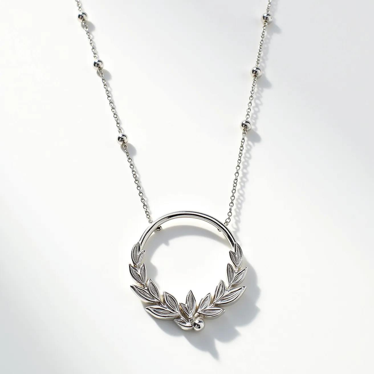 This white gold necklace features a delicate chain with small, polished beads evenly spaced along its length, adding a touch of elegance. At the center is a circular pendant, showcasing an intricate laurel wreath design, which is both classic and sophisticated. The laurel leaves are crafted in white gold, exhibiting fine detailing that enhances the piece's overall charm. The necklace likely uses a simple clasp mechanism for secure attachment, though the specific type of clasp is not visible. There are no visible gemstones set in the necklace, keeping the design minimalistic yet refined.