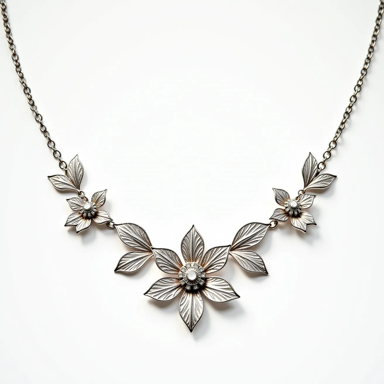 This white gold necklace features an intricate design with floral motifs, composed of finely detailed petals that branch out to form three distinct flowers. Each flower is accented with a small, round-cut diamond at its center, secured in a bezel setting that complements the elegance of the necklace. The petals radiate with a subtle texture, enhancing the reflective qualities of the white gold. The necklace links are crafted in a delicate chain that seamlessly connects each floral element. A discreet clasp secures the piece, ensuring both functionality and style, making it an exquisite example of jewelry artistry.