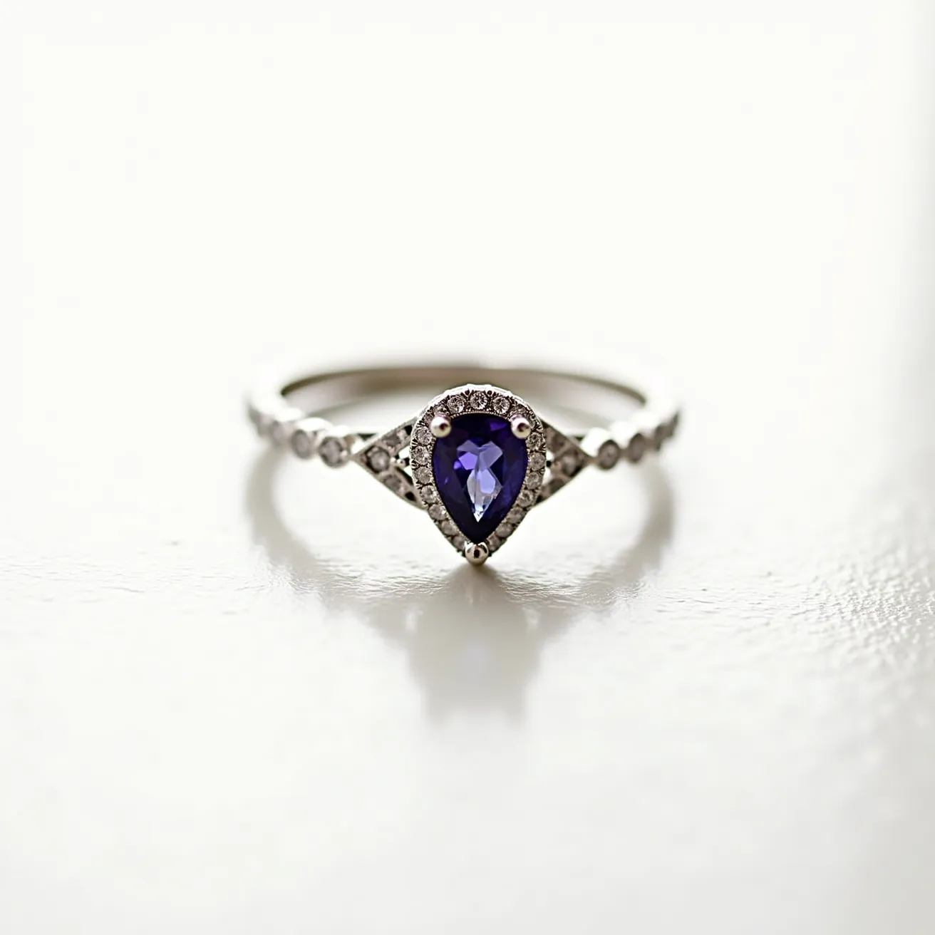 This alexandrite ring features a striking pear-shaped alexandrite stone set at its center, displaying its characteristic color-changing properties. The alexandrite is surrounded by a halo of small, round diamonds, enhancing its brilliance and adding a touch of elegance. The band itself appears to be crafted from white gold or platinum, matching the cool tones of the gemstones. On either side of the center setting, additional small diamonds accent the band, lending a refined sparkle to the overall design. The prong setting secures the alexandrite, ensuring it catches light from every angle.