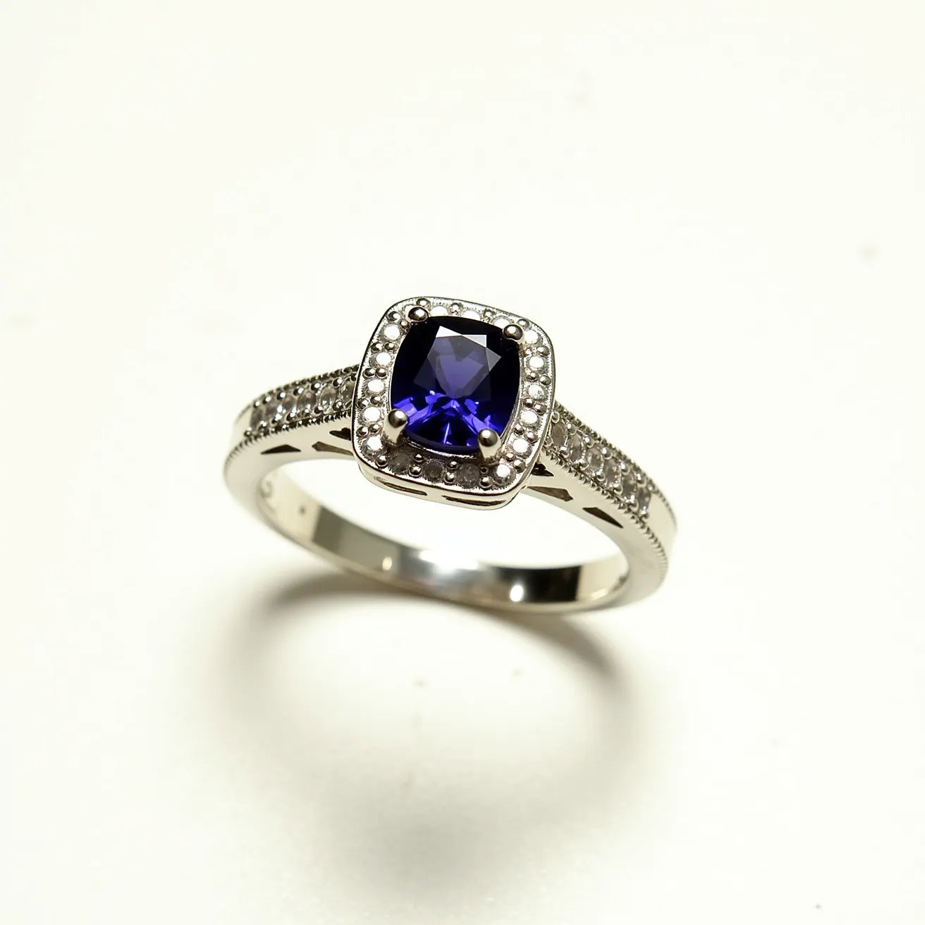 This alexandrite ring features a stunning cushion-cut central stone, known for its color-changing properties, set in a prong setting that highlights its brilliance. The metal band appears to be made of white gold or platinum, adding a complementary, cool-toned backdrop to the vibrant gemstone. Surrounding the alexandrite is a halo of small round diamonds, enhancing its allure with added sparkle and sophistication. The band itself is adorned with pavé-set diamonds, contributing to the ring's overall elegance and giving it an exquisite finish.