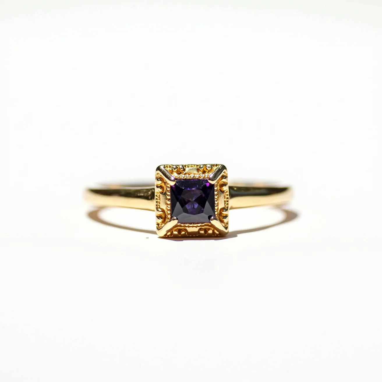 This alexandrite ring features a square-cut alexandrite gemstone set in an ornate gold setting. The gold band complements the stone, while the intricate details around the setting add elegance and highlight the central gem. The alexandrite's deep hues are accentuated by the precise cut, enhancing its visual appeal. Overall, the ring showcases a harmonious blend of luxury and craftsmanship.