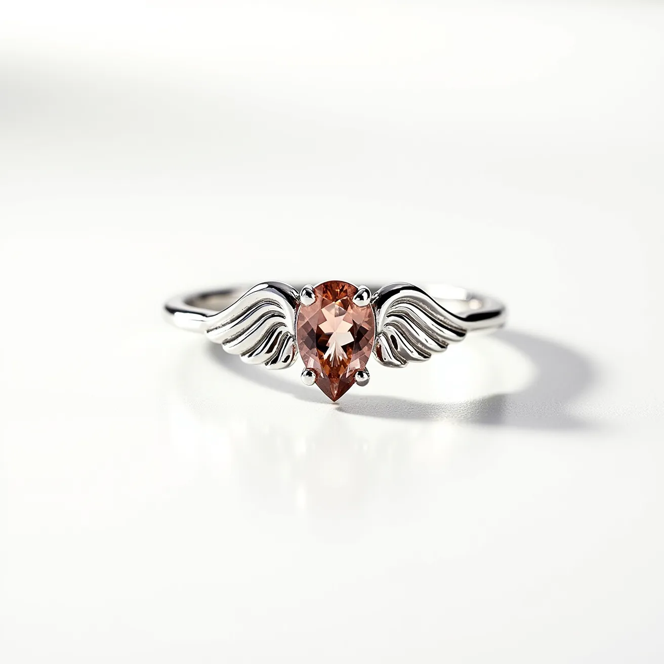 This angel ring features a sleek metal band crafted from a shiny silver material. At its center is a pear-shaped orange gemstone, securely held in place by a prong setting. The gemstone is flanked by delicate angel wing motifs on either side, intricately designed to enhance the celestial theme of the piece. The wings seamlessly integrate into the band, giving the ring a harmonious and elegant look, with no visible clasps or attachments disrupting its smooth design.