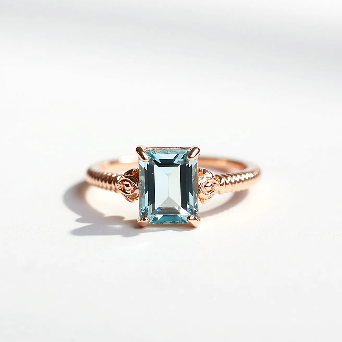 This aquamarine ring features a prominent rectangular-cut aquamarine gemstone, showcasing a light blue hue that captures and reflects light beautifully. The gem is securely set in a four-prong setting, highlighting its clean lines and facets. The band is likely crafted from rose gold, with intricate detailing that includes a twisted design, adding a touch of elegance and sophistication to the overall appearance. The ring’s setting and design elements work harmoniously to accentuate the central gemstone, creating a striking and stylish piece of jewelry.