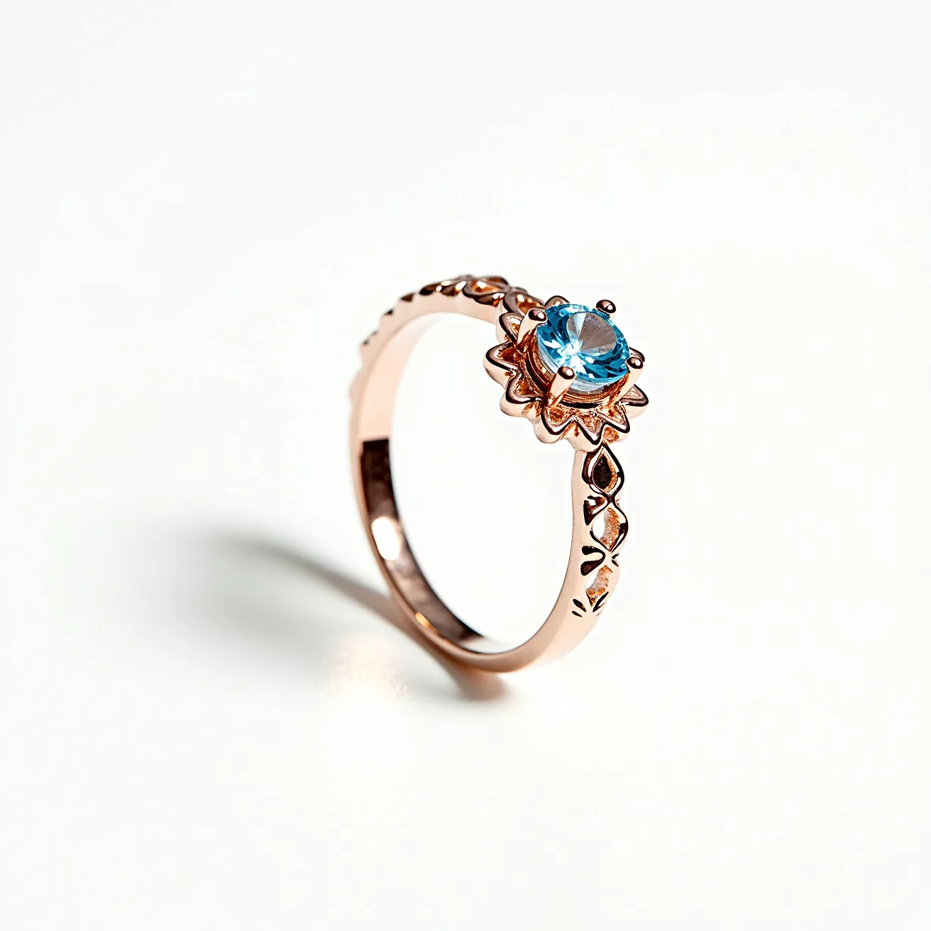 This aquamarine ring features a prominent round-cut aquamarine gemstone set in a rose gold band. The gem is securely placed in a decorative prong setting with a flower-like design that enhances its elegant appearance. The band itself is intricately detailed with a series of cut-out patterns that add to the ring’s delicate aesthetics. The combination of the cool blue aquamarine and the warm tone of the rose gold creates a striking contrast, making it a visually appealing piece.