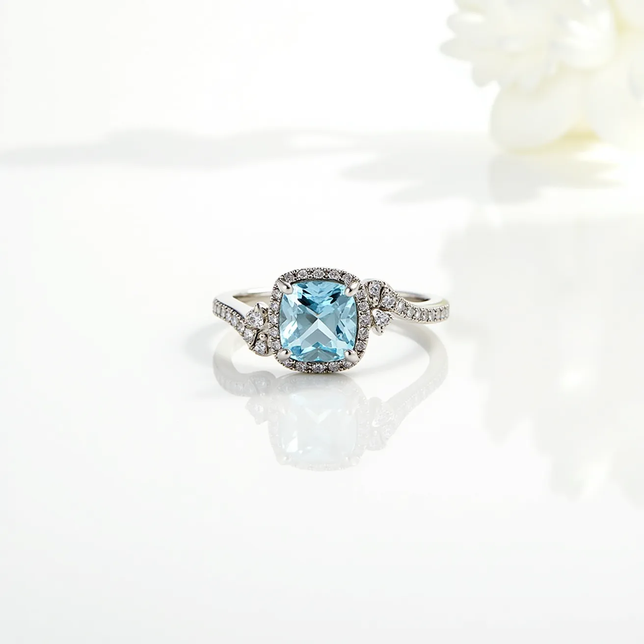 This aquamarine ring features a cushion-cut aquamarine stone as its centerpiece, set in a halo setting surrounded by small diamonds. The band is made of what appears to be white gold or platinum, enhancing the cool tones of the aquamarine and surrounding diamonds. The band itself is set with additional small diamonds, creating a flowing, intricate design that complements the central gem. The ring showcases a classic yet elegant design, with detailed craftsmanship evident in the meticulous setting of the stones. The combination of aquamarine and diamond accents makes this piece a standout for its sparkle and sophisticated allure.