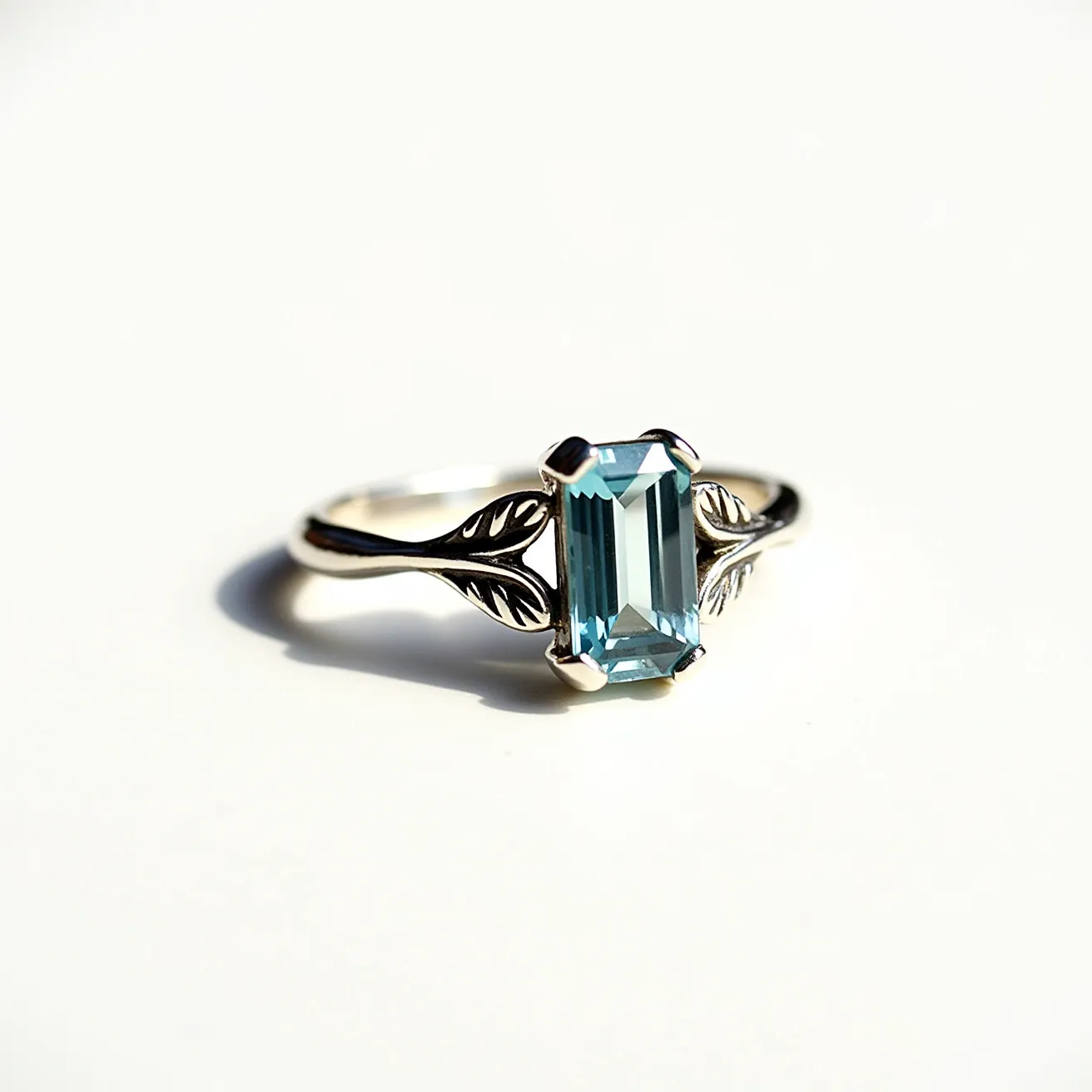 This aquamarine ring features a stunning, rectangular emerald-cut aquamarine gemstone set as the centerpiece. The gemstone is held securely by a four-prong setting, highlighting its clarity and icy blue hue. The band, likely made of polished white gold or platinum, is intricately designed with leaf-like motifs on either side of the stone, adding an elegant and naturalistic touch to the piece. The combination of the cool blue aquamarine with the bright metal setting creates a sophisticated and timeless appearance.