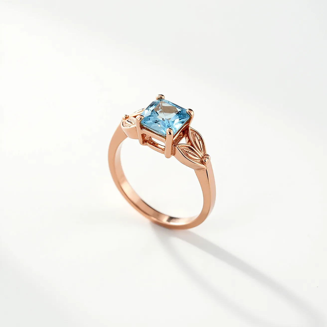 This aquamarine ring features a central square-cut aquamarine gemstone set in a prong setting, enhancing its visibility and providing structural stability. The stone exhibits a light blue hue characteristic of aquamarine. The band of the ring is crafted from a metal with a rose-gold finish, offering a warm contrast to the cool tones of the aquamarine. The design includes delicate leaf motifs on either side of the central stone, adding a touch of elegance and detail to the ring's overall aesthetic.