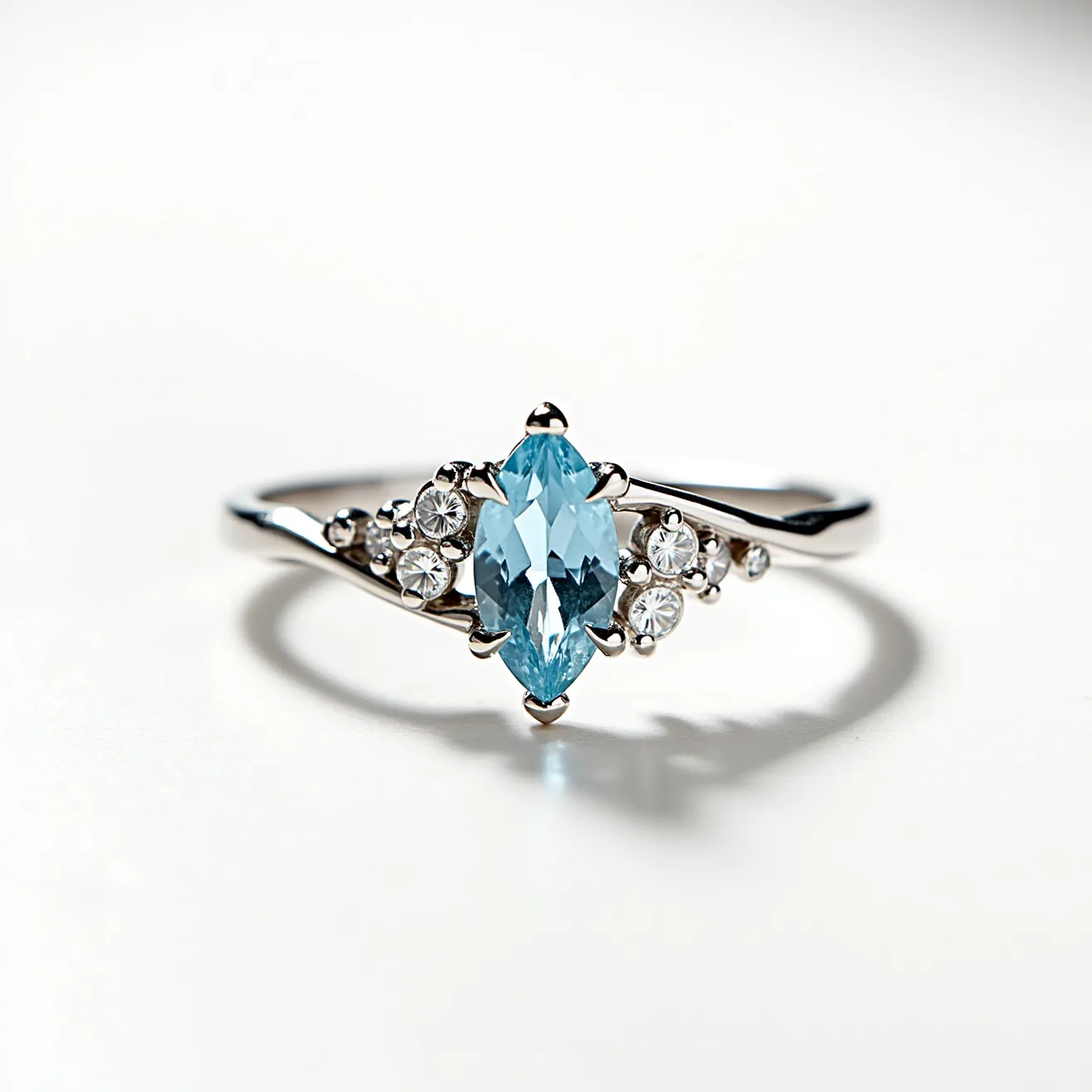 This aquamarine ring features a beautifully cut marquise aquamarine stone set in a sleek metal band, likely white gold or platinum. The central aquamarine stone is prong-set, allowing for maximum visibility and light reflection, enhancing its sparkling blue hue. Surrounding the aquamarine are smaller round-cut stones, resembling diamonds, which are set in a cluster formation on either side of the main stone, adding an elegant and sophisticated touch to the overall design. The smooth, polished band curves gracefully, complementing the classic yet modern aesthetic of the ring.