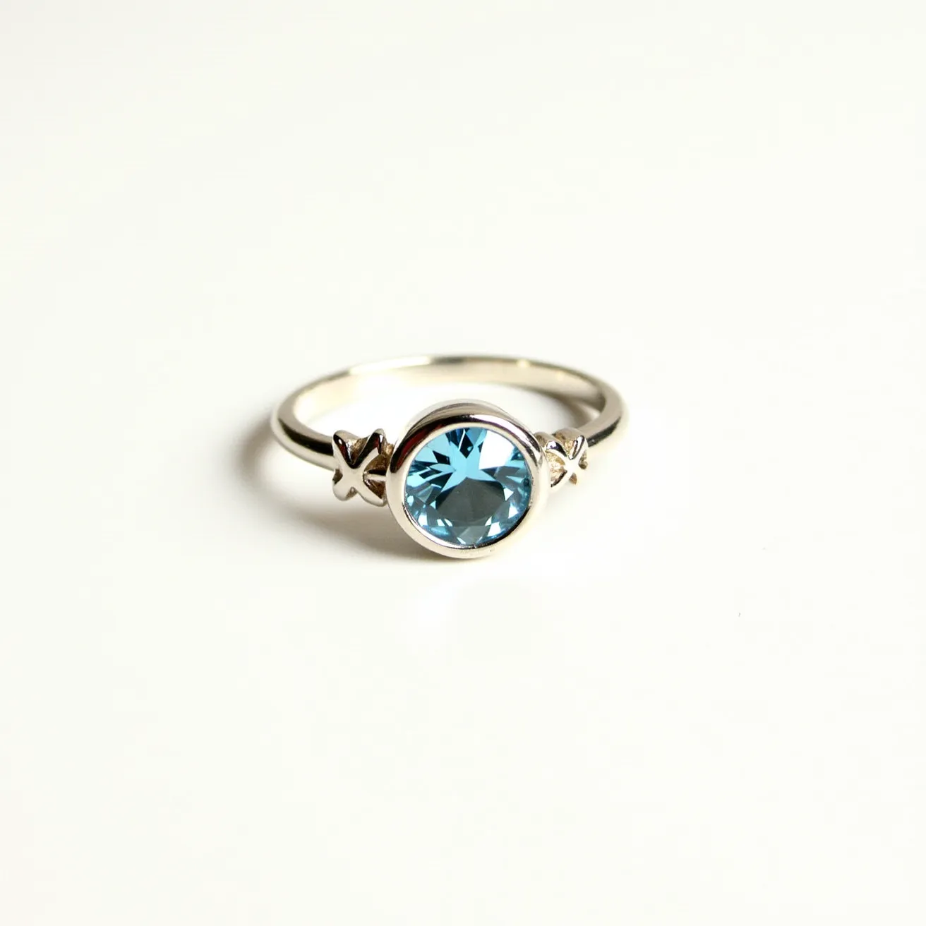 This aquamarine ring features a round-cut aquamarine stone set in a sleek bezel setting, showcasing its brilliant blue hue. Flanking the gemstone are two decorative elements that resemble small crosses, likely crafted from the same material as the band, which appears to be polished silver or white gold. The band itself is smooth and simple, highlighting the central stone and its unique design details without any additional clasps or attachments.