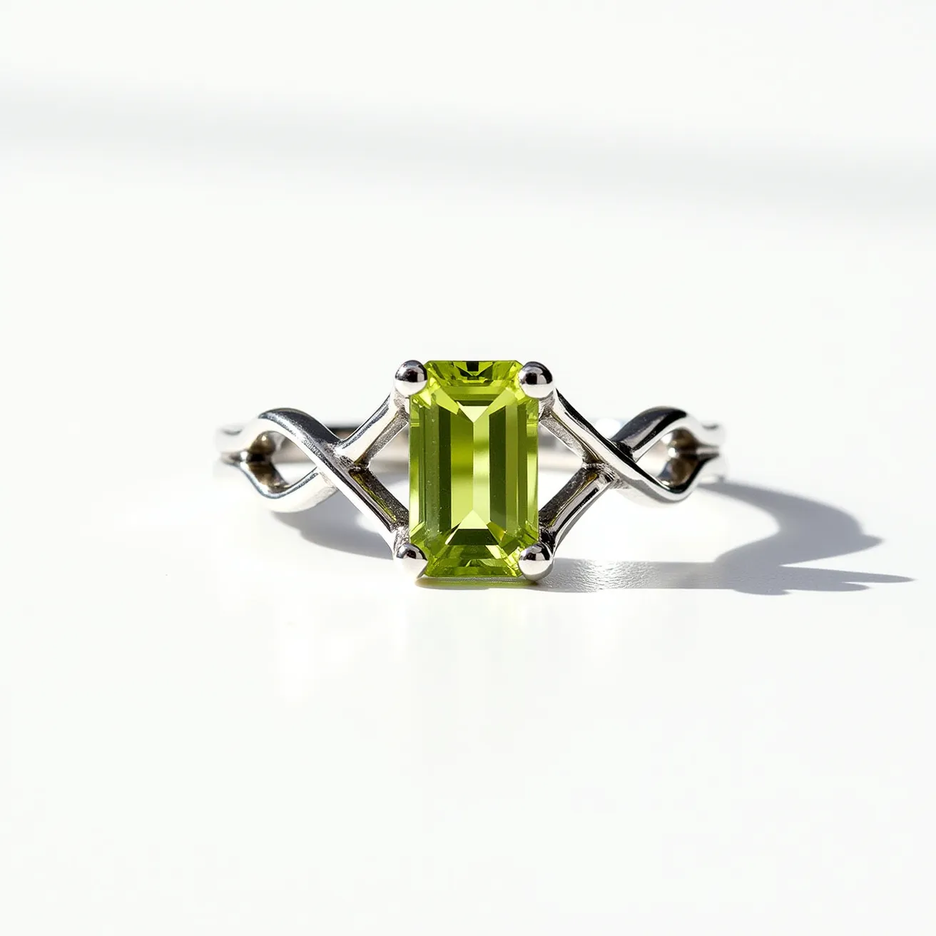 This august birthstone ring features a striking peridot, cut in an emerald shape, which is known for its vivid green hue. The peridot is prominently set in a four-prong setting, ensuring both security and elegance. The band of the ring is crafted from a polished metal, likely sterling silver or white gold, and displays a delicate intertwined design on each side of the stone, adding to the ring's sophisticated and modern appeal.