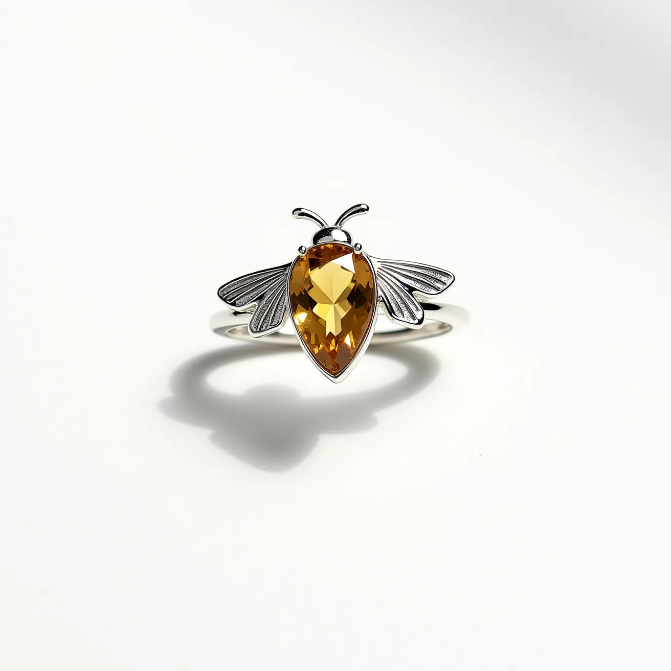 This bee ring features a striking yellow gemstone at its center, cut into a pear shape, which forms the body of the bee. The stone is secured with a prong setting and exhibits a brilliant facet arrangement, emphasizing its vibrant hue. The ring itself appears to be crafted from a polished metal, likely silver, forming the band and detailing of the bee's wings and antennae. The wings are intricately designed, showcasing delicate, engraved lines that add a realistic element to this charming piece. The overall design of the ring captures the essence of a bee with elegance and craftsmanship.