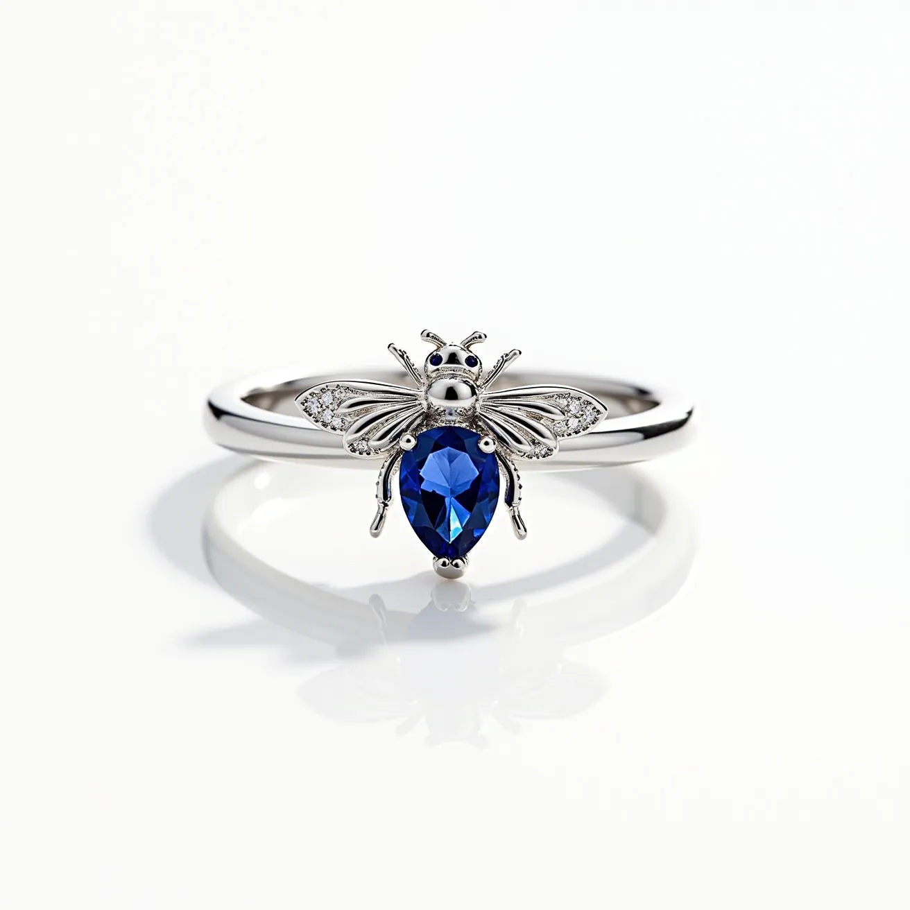 This bee ring features an elegant design crafted in metal that appears to be silver-toned. The centerpiece of the ring is a bee motif adorned with a sizable pear-shaped blue gemstone, set securely at the body of the bee. The wings are embellished with small, sparkling stones that are likely diamonds or diamond-like in appearance, adding a touch of brilliance to the piece. The ring band itself is sleek and polished, seamlessly integrating with the bee design, enhancing the overall aesthetic of this striking piece.