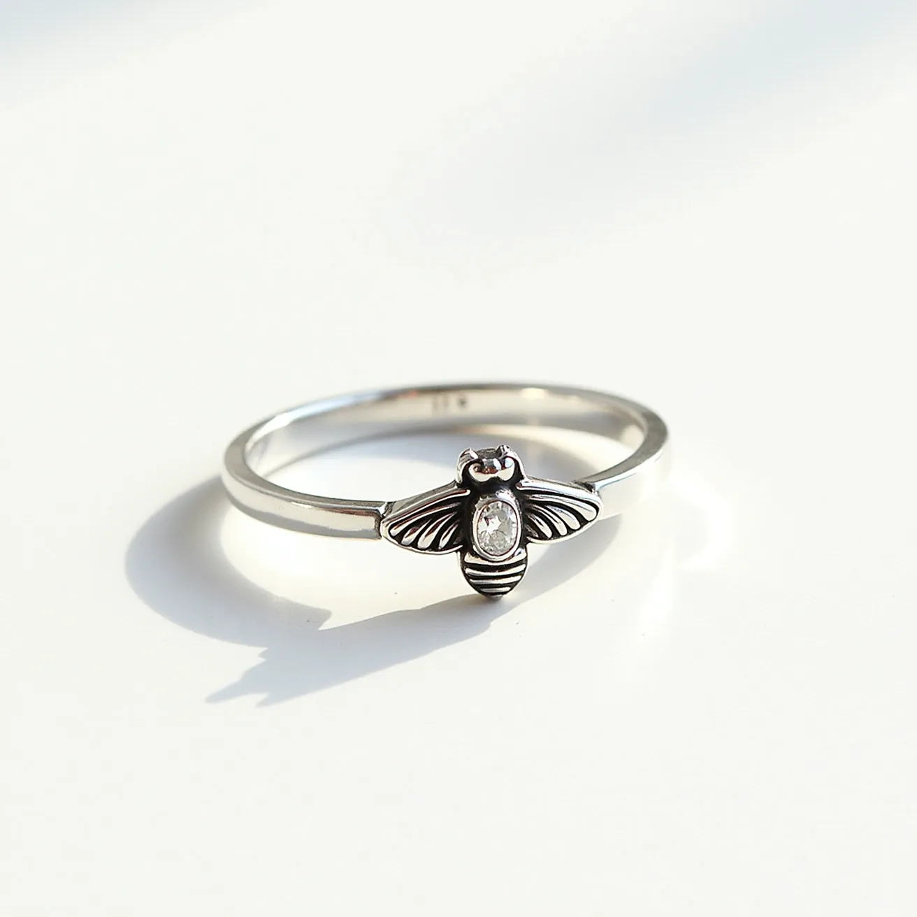 This bee ring features a sleek design crafted from what appears to be a polished silver material. At its center, there is a charming depiction of a bee, beautifully detailed with distinct wings and stripes. The bee is adorned with a small, oval-cut gemstone set in the middle, likely a diamond or a similar clear stone, adding a touch of elegance and sparkle to the piece. The stone is secured in a bezel setting, which complements the minimalist style of the ring. The band is smooth and thin, making it a delicate and stylish accessory.