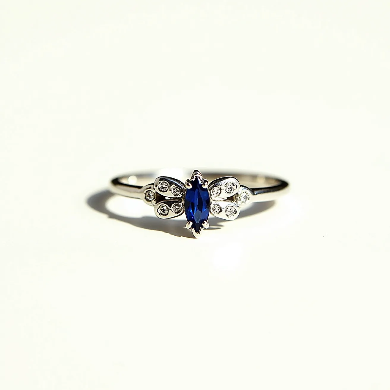 This bee ring features a polished metal band, likely silver or white gold, designed to resemble a bee. At its center, there is a marquise-cut sapphire, serving as the body of the bee, set in a prong setting for security and visibility. Flanking the sapphire on either side are small round brilliant-cut diamonds arranged to suggest wings, adding a touch of sparkle and dimension. The detailed craftsmanship highlights a combination of elegance and whimsy, capturing the essence of a bee in flight. The ring's band is sleek and subtly tapers towards the setting, enhancing both comfort and aesthetic appeal.