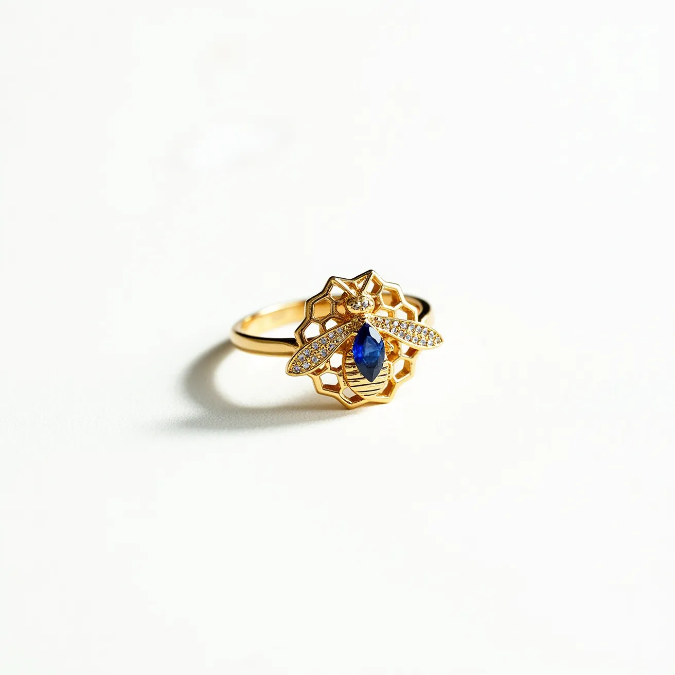 This bee ring features a detailed bee design, crafted in a gold-tone material, sitting atop an intricate honeycomb structure. The body of the bee is adorned with a striking marquise-cut blue gemstone, set prominently to stand out. The bee's wings are elegantly embellished with a series of small, sparkling diamonds or diamond-like stones, adding to the overall shimmer and sophistication of the piece. The ring's band is simple, allowing the ornate bee and honeycomb centerpiece to capture full attention. The combination of metals and gemstones creates a luxurious and eye-catching accessory.