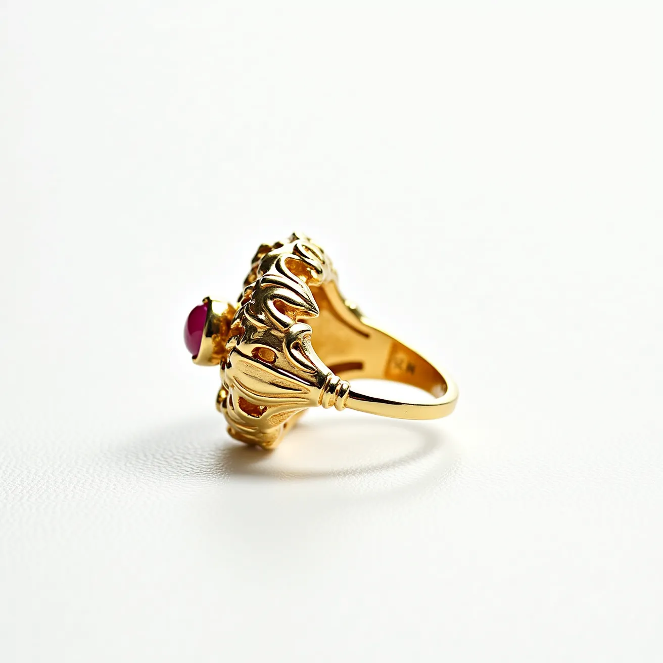 This big ring features an intricate gold setting with ornate detailing, showcasing its craftsmanship. At the center, a striking red gemstone, likely a cabochon-cut ruby, is securely set in a crown-like bezel. The gold band is smooth and polished, complementing the elaborate design of the upper structure, which has a decorative, almost floral appearance. The combination of the red gem and the warm gold metal gives the ring a regal aesthetic, making it a statement piece of fine jewelry.