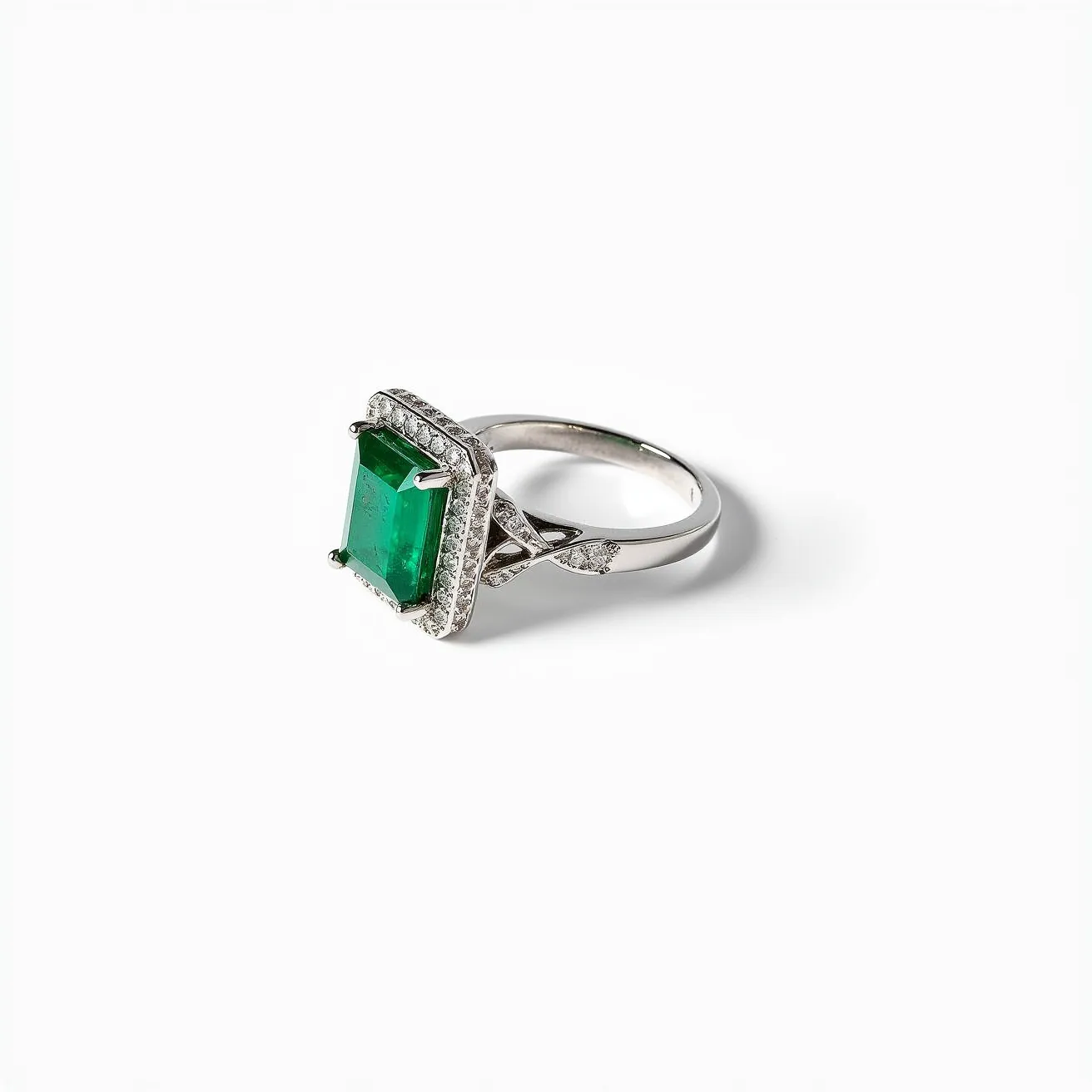 This big ring features a prominent emerald-cut green gem at its center, securely held by four prongs. The large stone is showcased within a halo of smaller, round-cut clear stones, which enhance its vibrant color and sparkle. The ring is crafted from a lustrous metal, possibly platinum or white gold, lending it an elegant and timeless appeal. The band incorporates intricate latticework near the setting, adding further detail and sophistication to the design. The overall composition of the ring reflects a classic yet luxurious style.