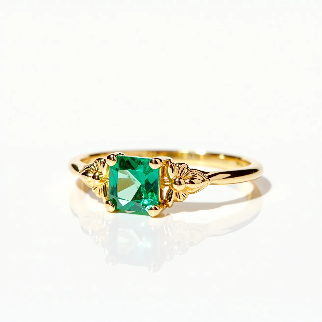 This big ring features a striking square-cut green gemstone, likely an emerald, set prominently at its center. The stone is secured in place by a delicate prong setting, crafted from polished yellow gold. Flanking the gemstone on either side are intricate floral motifs that add decorative detail to the ring's band. The gold shank of the ring is smooth and gleaming, creating a sophisticated contrast to the vibrant green of the gemstone. The design of the ring reflects both elegance and a touch of vintage charm, making it a standout piece.