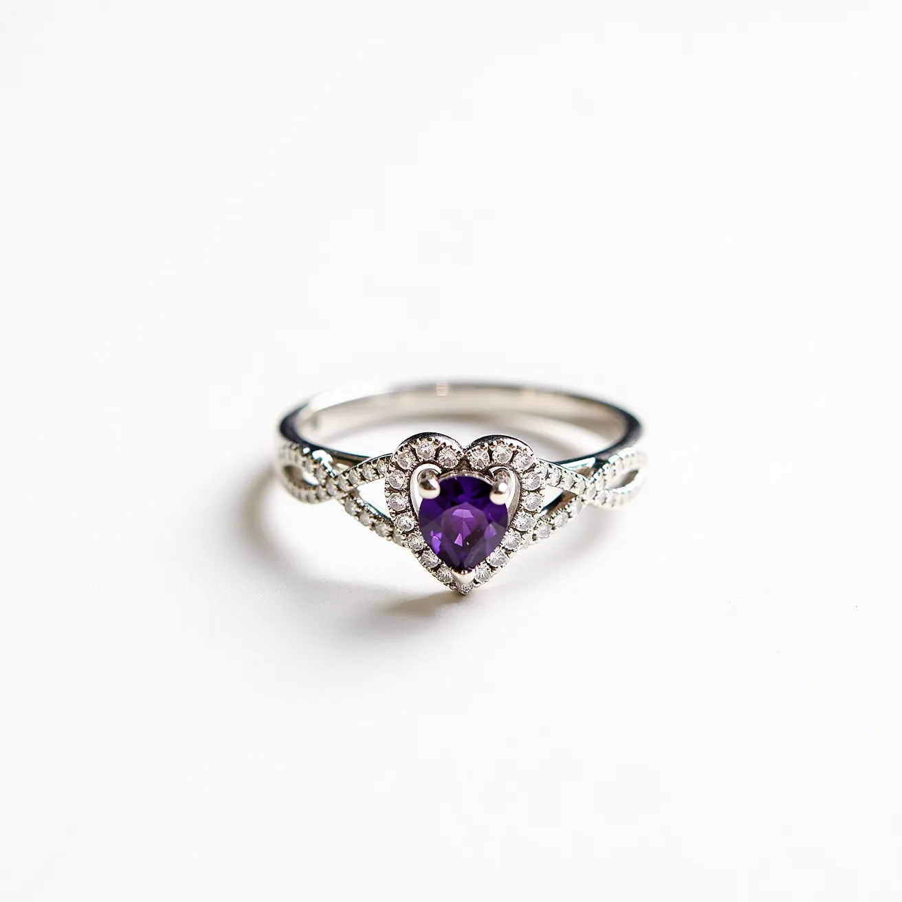 This birthstone ring features a central heart-shaped amethyst gemstone, notable for its vibrant purple hue, set in a secure prong setting. The band is crafted from silver, adding to its polished and elegant appearance, and is intricately designed with small round diamonds or diamond-like stones that embellish the surrounding area, enhancing the ring's overall sparkle. The stones are arranged in a graceful, crisscrossing pattern on the band, which adds a sophisticated touch to its design.