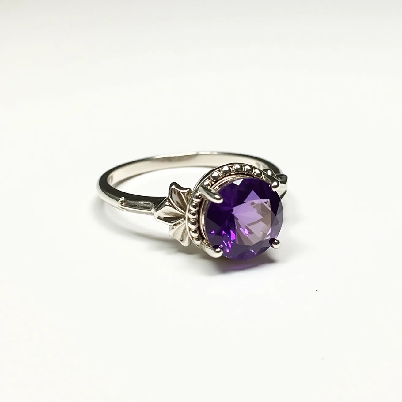 This birthstone ring features a central round-cut amethyst, which is securely set in a bezel with a raised border adorned with intricate detailing. The amethyst, displaying a rich purple hue, is highlighted by its polished setting, which appears to be crafted from a silvery metal, possibly sterling silver. The ring’s band is slender and smooth, providing a graceful setting for the striking gemstone. The overall design combines elegance with a touch of vintage charm, making it a lovely piece for any occasion.