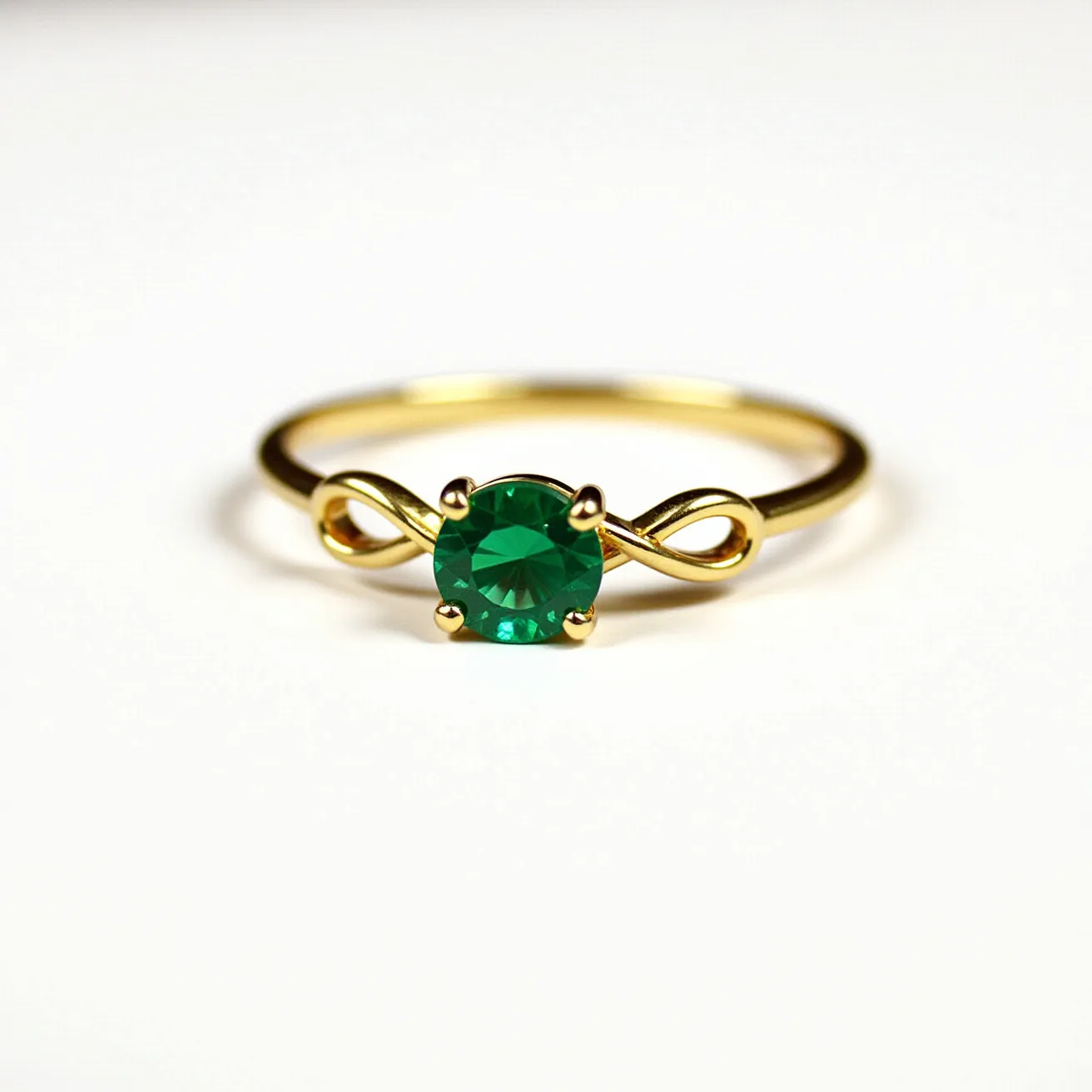 This birthstone ring features a striking emerald-green gemstone at its center, cut in a round shape which captures and reflects light beautifully. The stone is set in a classic four-prong setting, ensuring its security while highlighting its brilliance. The band appears to be crafted from a polished gold material, adding a touch of warmth and elegance. An intricate infinity design forms the shoulders of the ring, seamlessly connecting the band to the stone, symbolizing timelessness and continuity.