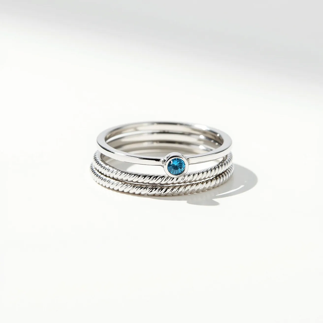 These birthstone stackable rings feature a set of sleek, polished bands with a bezel-set blue gemstone at the center, likely representing a birthstone. The primary materials of the rings seem to be silver or a silver-toned metal, giving them a shiny, reflective quality. The central gemstone is round-cut, adding a touch of elegance and color contrast to the minimalist design. Some of the rings have a twisted rope design, adding texture and visual interest. There are no visible clasps or attachments, as these are slip-on style rings designed for stacking together on a finger.