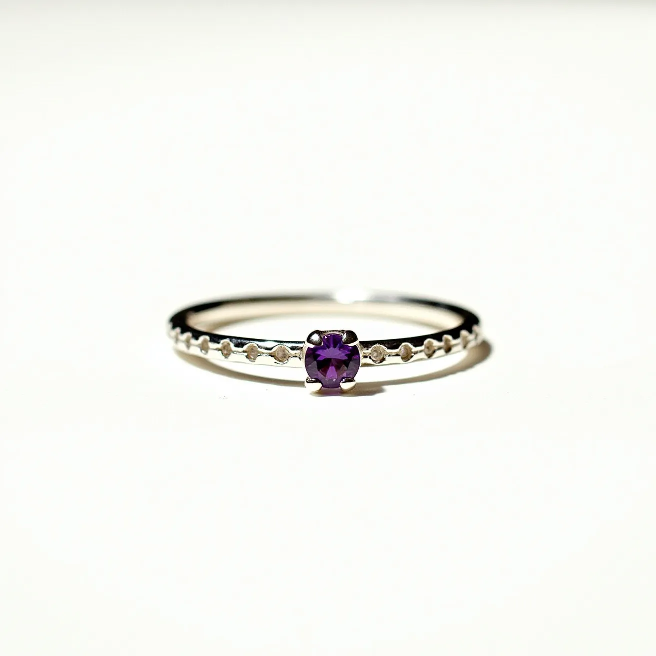 This birthstone stackable ring showcases a delicate design featuring a silver band, which is adorned with small, circular embellishments along its circumference. The focal point of the ring is a round-cut, vibrant purple gemstone, likely representing an amethyst birthstone. The gemstone is set securely in a classic prong setting, which enhances its visibility and sparkle. This elegant design allows for stacking with other rings, offering a versatile and personalized piece of jewelry.