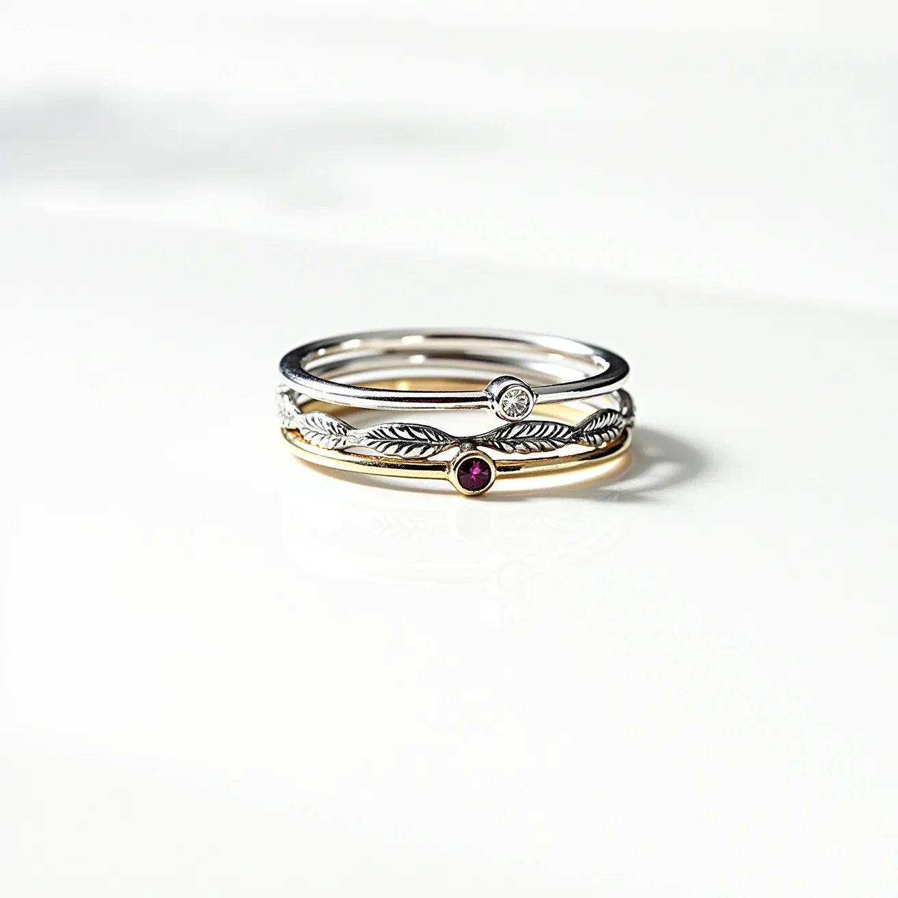 These birthstone stackable rings feature a set of three delicately crafted bands. The materials appear to be a combination of silver and gold tones, possibly sterling silver and gold plating. The topmost ring is a simple silver band with a small, clear round-cut gemstone, bezel-set as its centerpiece. The middle ring showcases a nature-inspired design, with intricate leaf motifs along the band, and is highlighted by a small white gemstone, also bezel-set. The bottom ring, in a warm gold tone, boasts a tiny, round, purple gemstone that stands out vividly against the band, similarly bezel-set. These rings are designed to stack seamlessly together, offering an elegant and personalized touch to the wearer's hand.