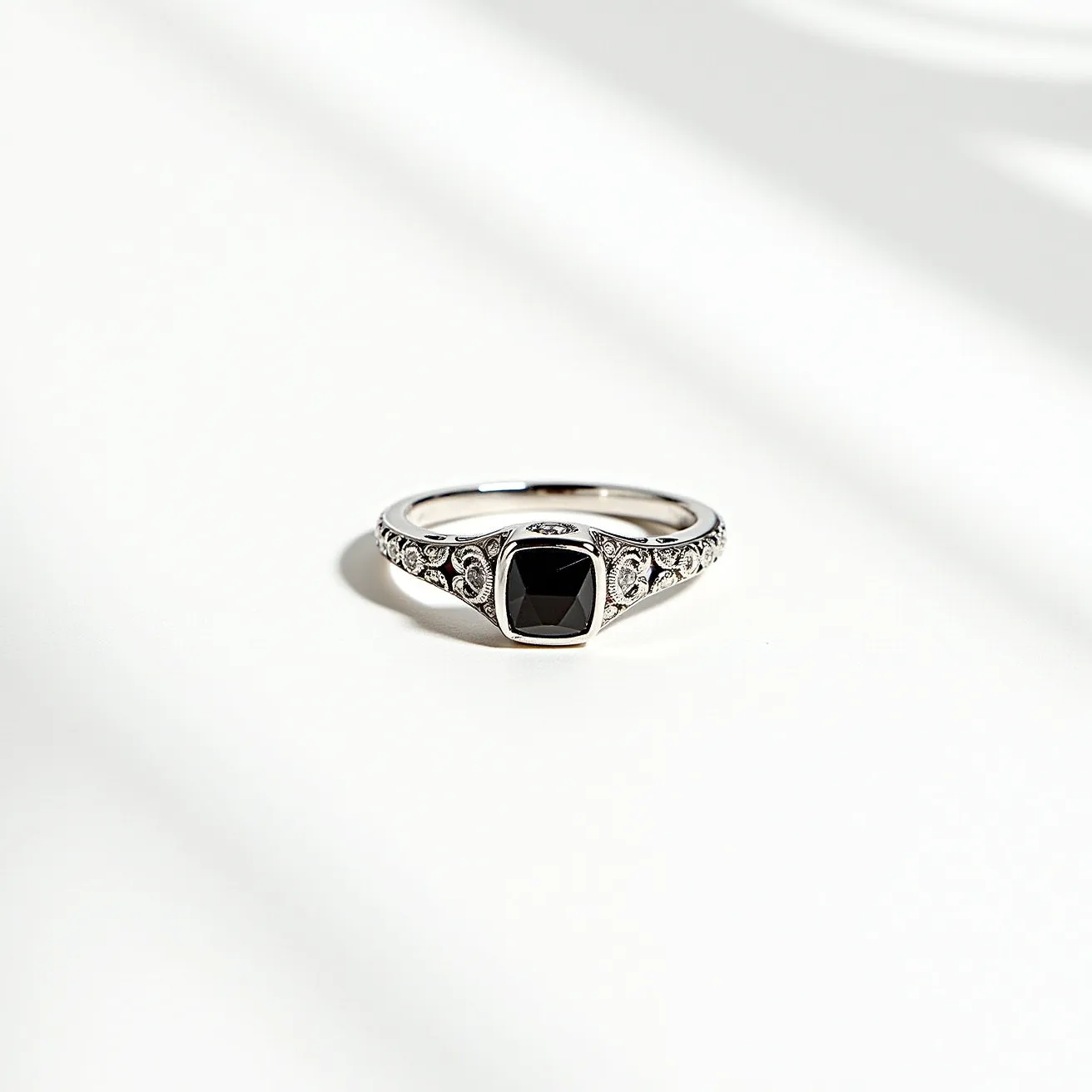 This black diamond ring features a striking square-cut black diamond set in a polished white metal band, likely white gold or platinum. The setting is a bezel, securely framing the gemstone and enhancing its modern aesthetic. The band is intricately engraved with decorative motifs, adding a touch of vintage elegance to the piece. Small, clear gemstones, possibly diamonds, are embedded into the detailed design on both sides of the main stone, providing additional sparkle and visual interest. The craftsmanship showcases a harmonious blend of contemporary and classic design elements, making it a unique and elegant piece of jewelry.