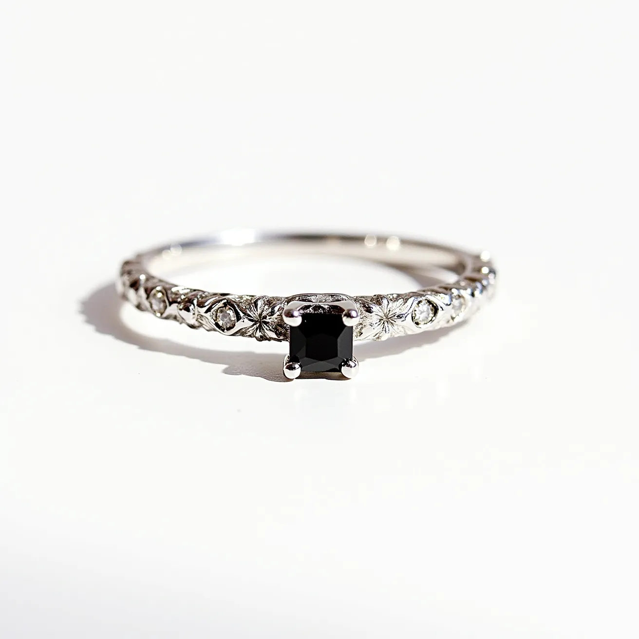 This black diamond ring features a striking square-cut black diamond set prominently in a prong setting, which secures the stone while allowing maximum light exposure. The band is crafted from a polished metal, likely white gold or platinum, and is intricately designed with floral or vine-like engravings that add an elegant touch. Small round brilliant-cut white diamonds are embedded along the band, complementing the central black diamond and enhancing the ring's sophisticated aesthetic. The overall design is both classic and modern, balancing the boldness of the black diamond with delicate detailing.