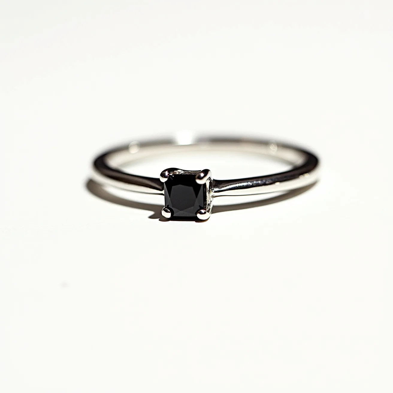 This black diamond ring features a solitaire black diamond at its center, presented in a minimalist design. The stone is cut in a square shape, likely a princess cut, and is securely set within a four-prong setting. The band of the ring appears to be made of a polished metal, possibly white gold or platinum, which complements the dark, opaque hue of the diamond. The overall design is sleek and modern, emphasizing the unique beauty of the black diamond. There are no additional stones or embellishments, highlighting the elegant simplicity of the piece.