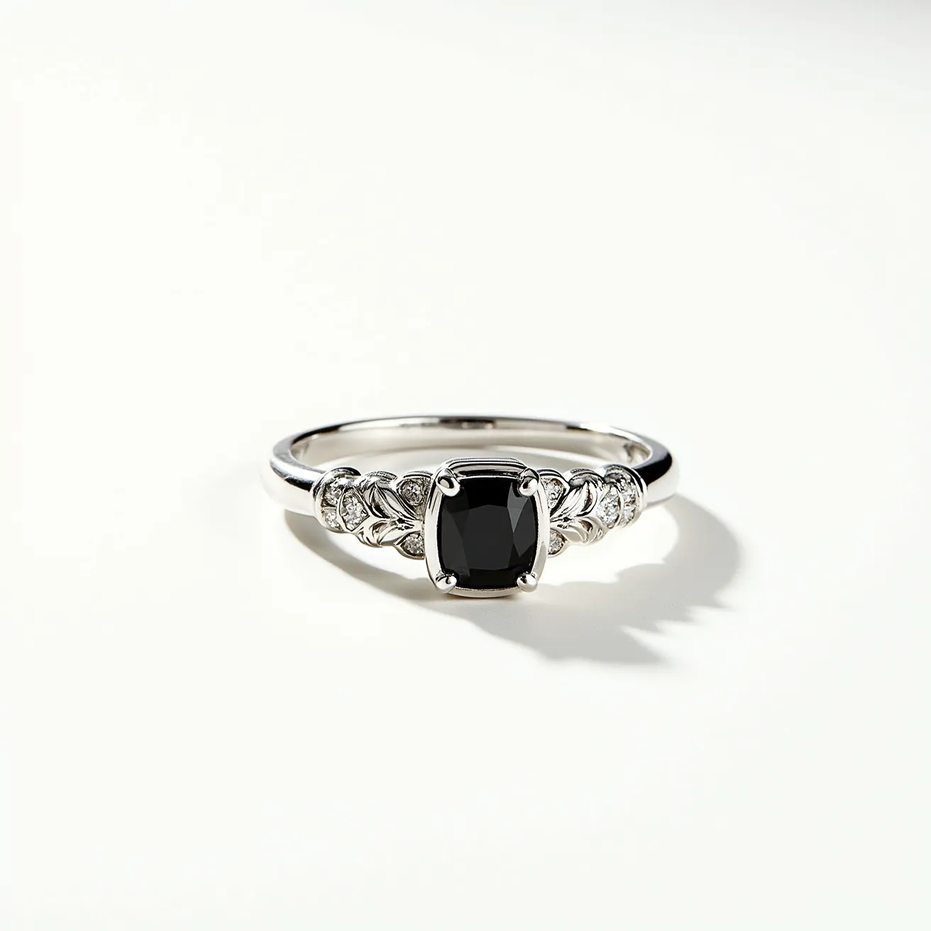 This black diamond ring features a cushion-cut black diamond set in a prong setting, surrounded by a delicate array of smaller diamonds embedded in the band. The band appears to be crafted from white gold or platinum, providing a sleek and elegant backdrop for the central stone. The arrangement of smaller diamonds forms a leaf-like design on either side of the centerpiece, enhancing the overall aesthetic with a touch of sophistication. The ring’s design emphasizes a balance between modern simplicity and intricate detailing, making it a striking piece of jewelry.