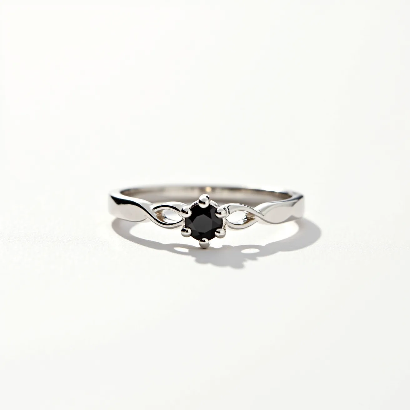 This black diamond ring features a sophisticated design with a central black diamond cut in a round shape, held securely in place by six prongs in a classic prong setting. The band appears to be crafted from a polished silver or white gold material, offering a sleek and reflective finish. The band showcases an elegant twisting design on either side of the diamond, adding a touch of modernity and complexity to the overall look. The prong setting allows for maximum visibility and light exposure to the diamond, enhancing the stone's subtle reflections and unique color.