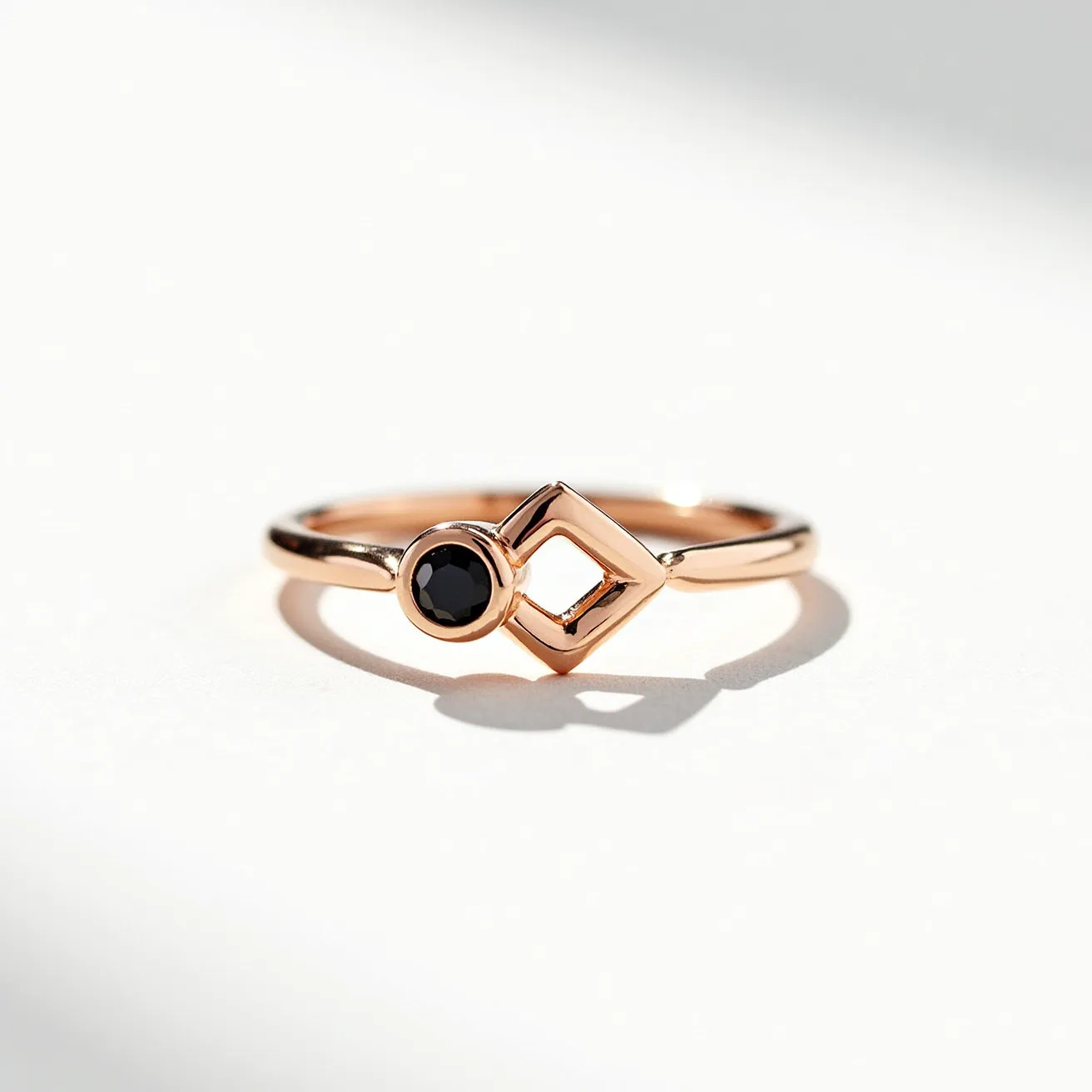 This black diamond ring features a sleek, polished band crafted from rose gold. The focal point is a round-cut black diamond, elegantly set in a bezel setting that enhances its dark, lustrous appearance. Adjacent to the gemstone, the design incorporates a geometric diamond-shaped motif, adding a modern and artistic element to the overall aesthetic. The combination of materials and design accents creates a unique and contemporary piece of jewelry. The band lacks any additional clasps, emphasizing a minimalist and seamless look.