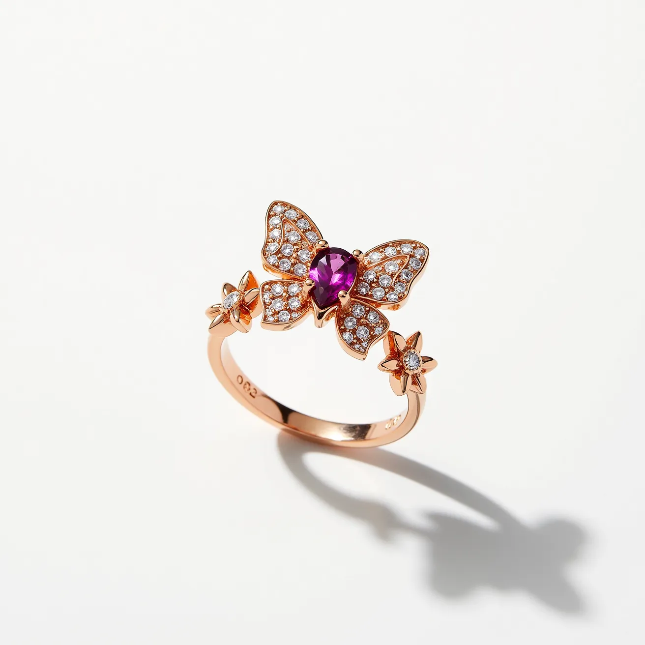 This butterfly ring features a beautifully crafted design in rose gold, showcasing a central butterfly motif adorned with a vibrant pink gemstone, likely a ruby or a similar stone, set in an oval cut at the center. Surrounding the central gem are numerous small, round-cut diamonds embedded into the wings of the butterfly in a pavé setting, enhancing its sparkle. Complementing the butterfly are two floral elements on either side of the band, each holding a small diamond at its center. The band itself is elegantly smooth, and the use of rose gold imparts a warm, luxurious glow to the piece.