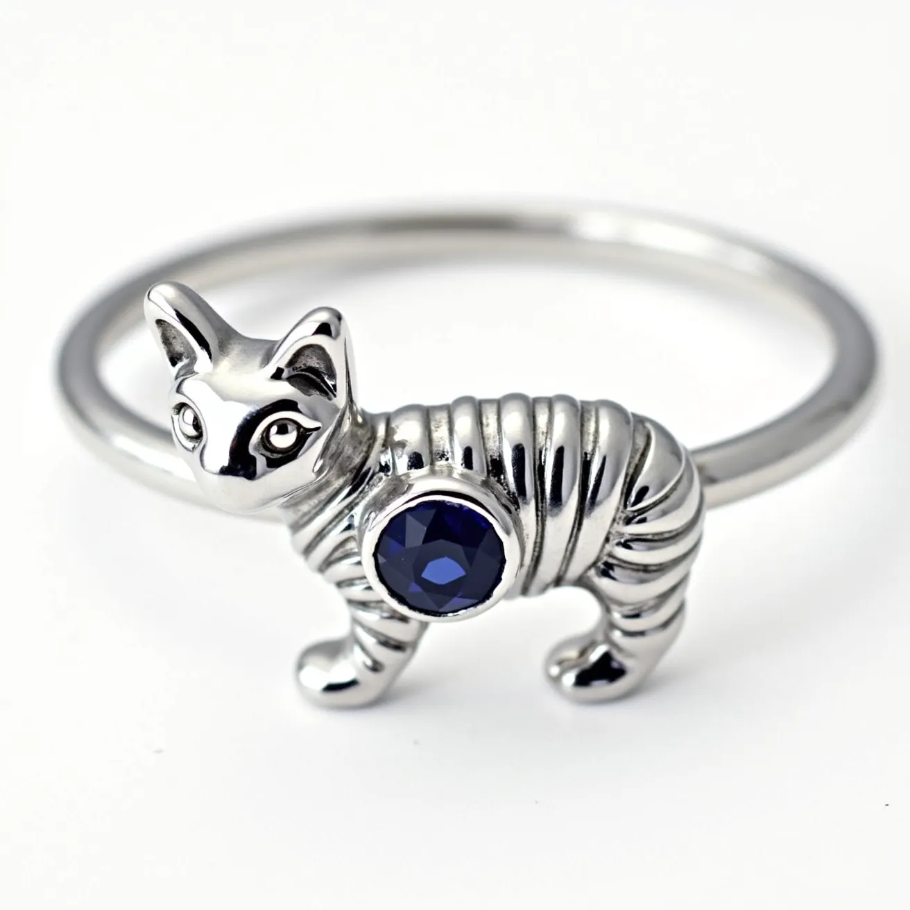 This cat ring features a sleek, silver-toned metal design shaped into a whimsical cat figure. The body of the cat is adorned with a round, deep blue gemstone, likely a sapphire or blue cubic zirconia, accentuating its playful elegance. The stone is set in a bezel setting, securely encased with smooth metal edges. The band is smooth and polished, seamlessly connecting to the cat figure without any visible clasps or attachments, creating a minimalistic and continuous design.