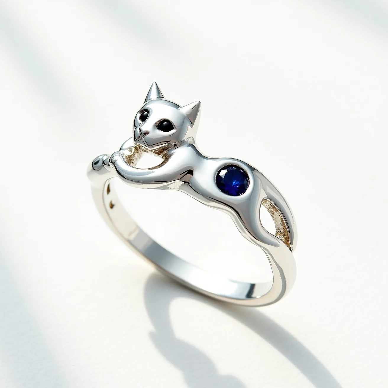 This cat ring is crafted from a shiny metal, likely silver or white gold, and features a distinctive design with a cat figure gracefully integrated into the band. The cat's eyes appear to be small black stones, adding a touch of character to its face. On the cat's body, there is a round, blue gemstone that is set in a smooth bezel setting, enhancing the gem's color and providing a secure hold. The band is smoothly polished, reflecting light and highlighting the detailed craftsmanship of the cat motif, which seamlessly flows into the ring's structure without any visible clasps or attachments.