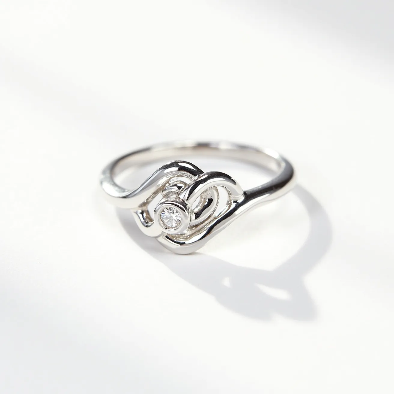 This chevron ring features a sleek, polished silver band that elegantly curves into a wave-like design. At its center, a round-cut diamond is nestled, securely held in a bezel setting that integrates seamlessly into the flowing lines of the ring. The simplicity of the metalwork enhances the brilliance of the diamond, creating a harmonious blend of modern and classic elements. The continuous band exhibits a fluidity that complements the central motif, lending an understated sophistication to the overall aesthetic.