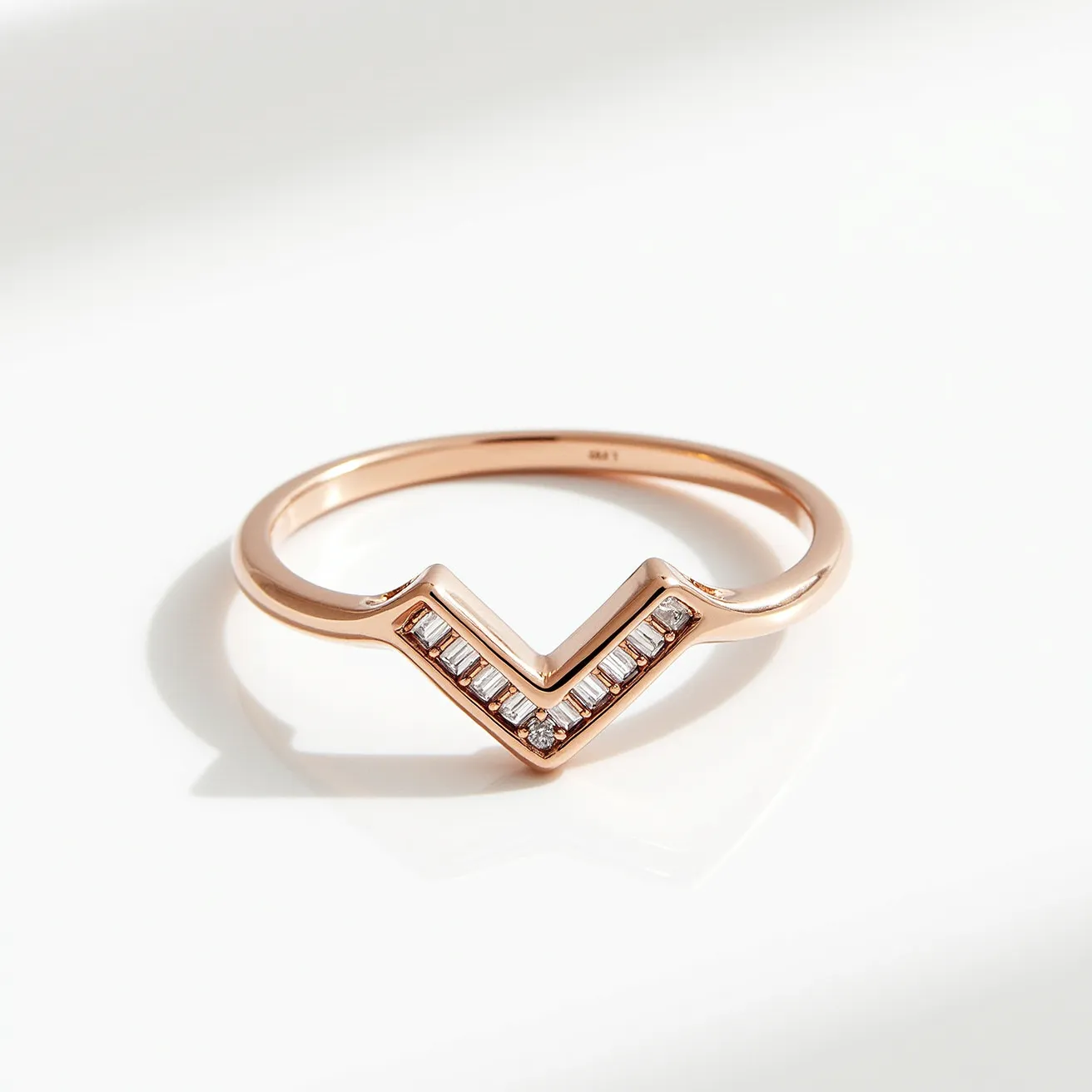 This chevron ring features a sleek and modern design crafted from a rose gold material, creating a warm and luxurious appearance. The ring is adorned with baguette-cut stones, most likely diamonds, neatly set into the V-shaped band. Each stone is positioned closely together in a channel setting, which provides a continuous line of sparkle along the chevron design. The simplicity of the band highlights the elegance and sharpness of the V-shape, allowing the stones to stand out. There is no visible clasp or attachment, indicating a continuous circular band designed for comfortable wearing.