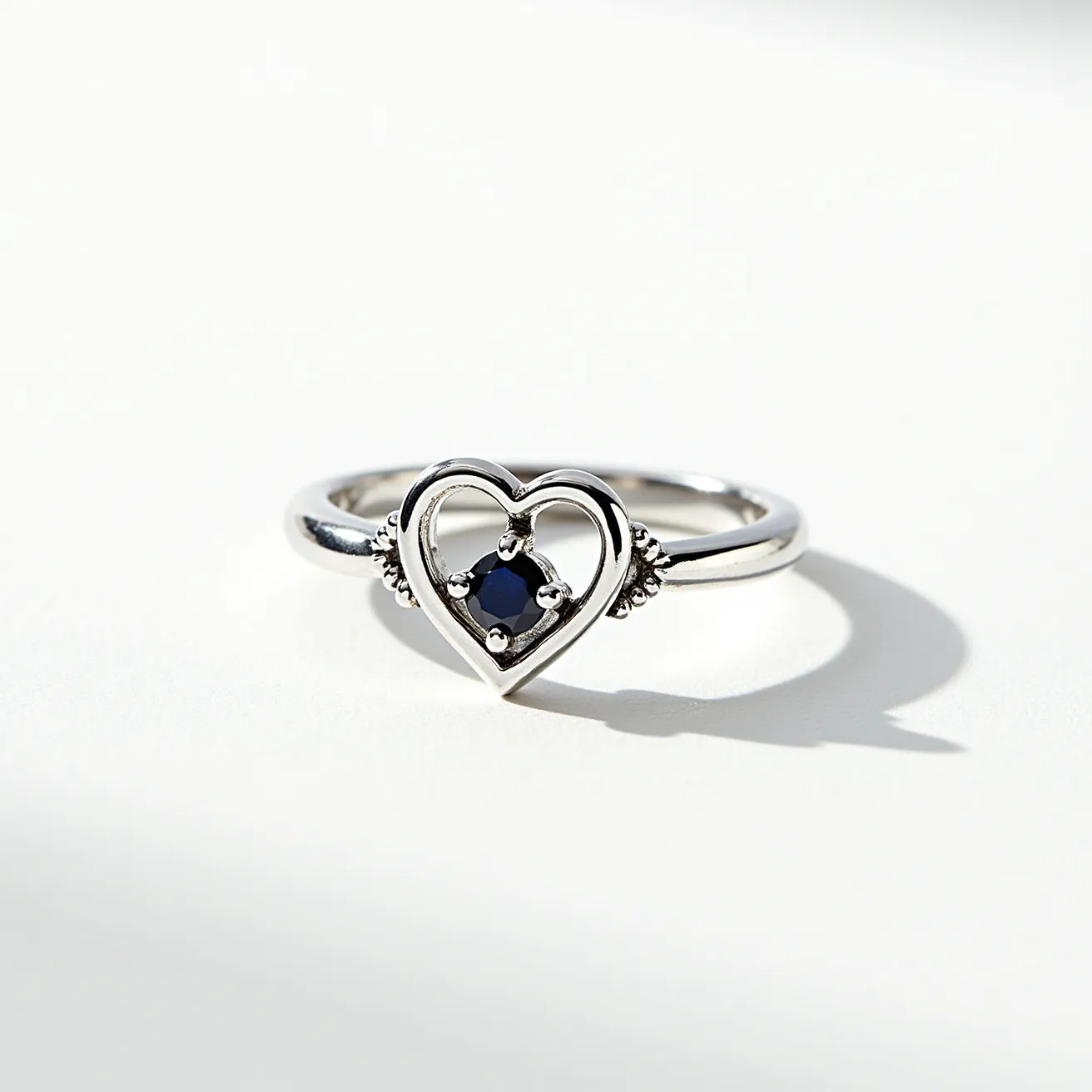 This Chrome Hearts ring features a sleek metal band, likely crafted from sterling silver, which is characteristic of the brand. At its center is an open-work heart motif, highlighting a single dark blue gemstone, probably a sapphire, set with four prongs, adding a touch of elegance and color contrast to the design. The prongs are evenly spaced, allowing the stone to be securely held in place, and small beaded details flank the sides of the heart, adding texture and visual interest to the piece. The open-heart design emphasizes simplicity and sophistication, making it a timeless accessory.