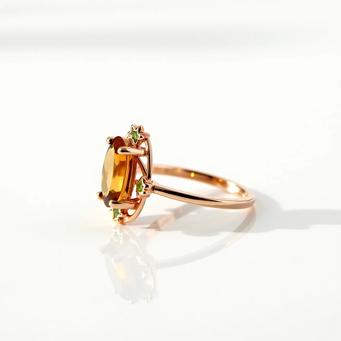 This citrine ring features a prominent oval-cut citrine stone as its centerpiece, set with prongs in a delicate rose gold band. The citrine displays a vibrant yellow-orange hue that is complemented by smaller, round green stones, likely peridots, set alongside the main stone, adding contrast and elegance to the design. The ring's band is smooth and slender, enhancing the visibility of the stones without any additional clasps or attachments, creating a refined and sophisticated appearance.