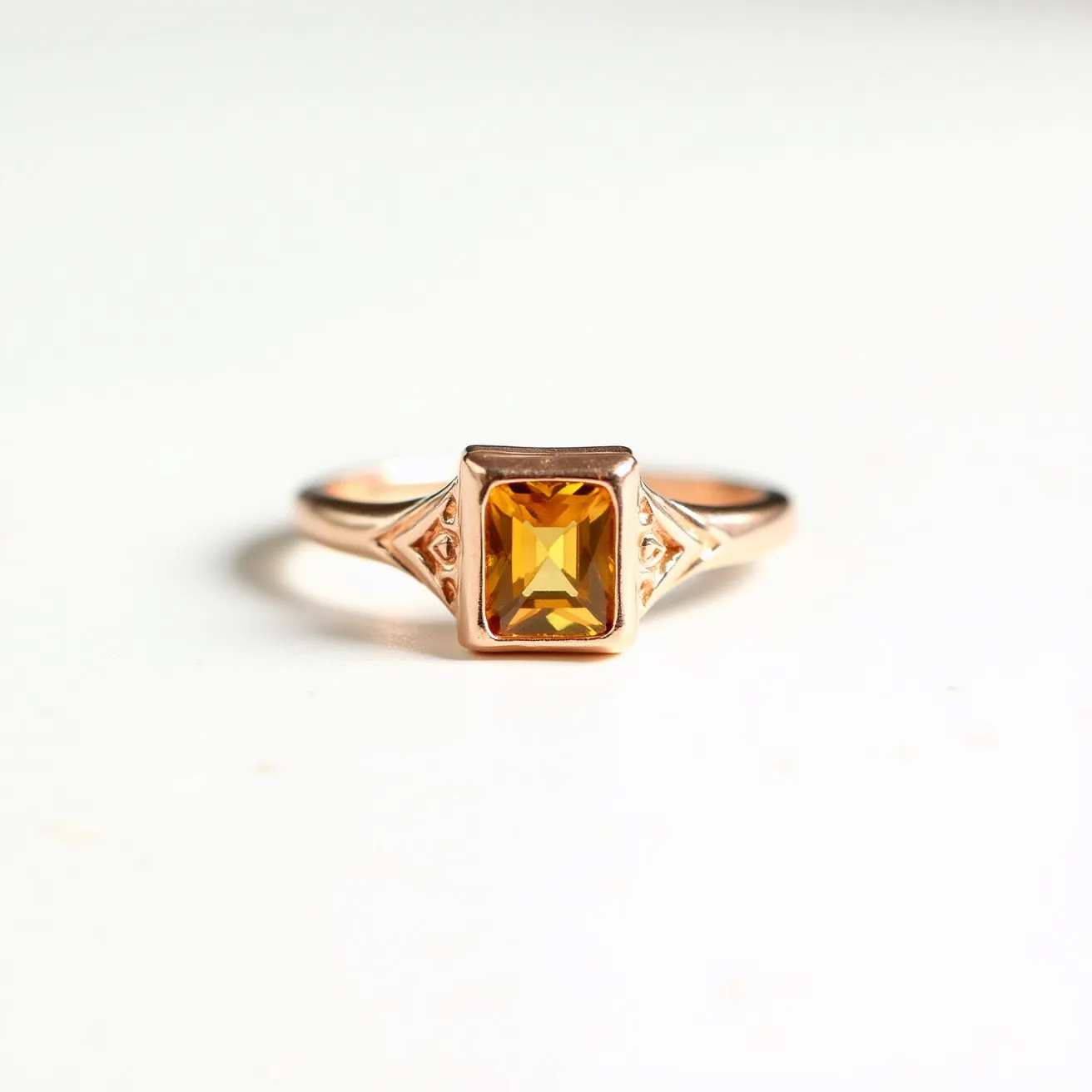 This citrine ring features a rectangular, emerald-cut citrine stone set in a sleek bezel setting. The ring is crafted from a polished metal, likely rose gold, complementing the warm hues of the citrine. The design is enhanced with two small decorative elements on each side of the stone, adding elegance to the overall setting. The band has a smooth finish, providing a comfortable fit.